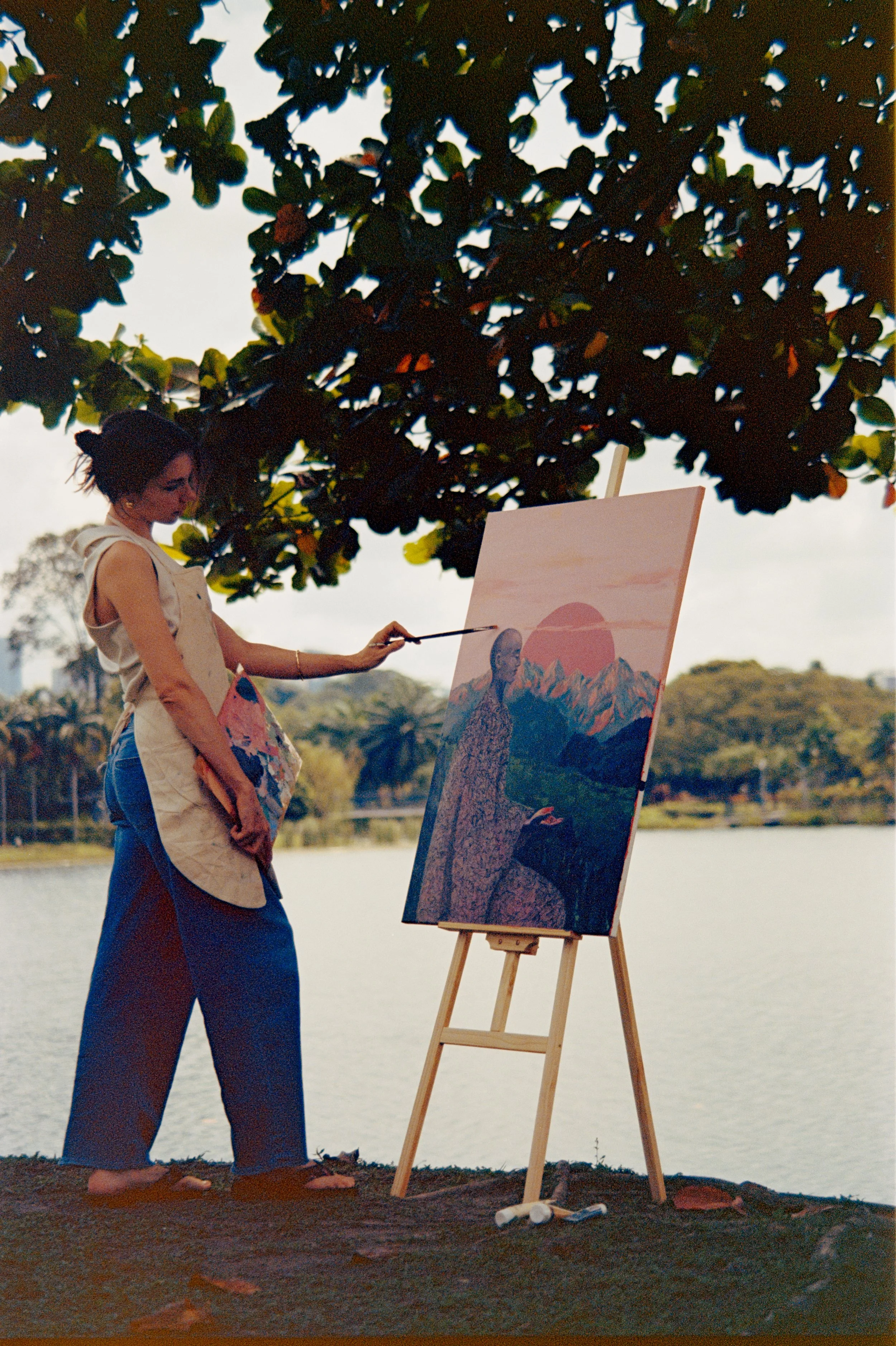 Woman painting a landscape on an easel outdoors by a lake, with trees and mountains in the distance under an overcast sky.