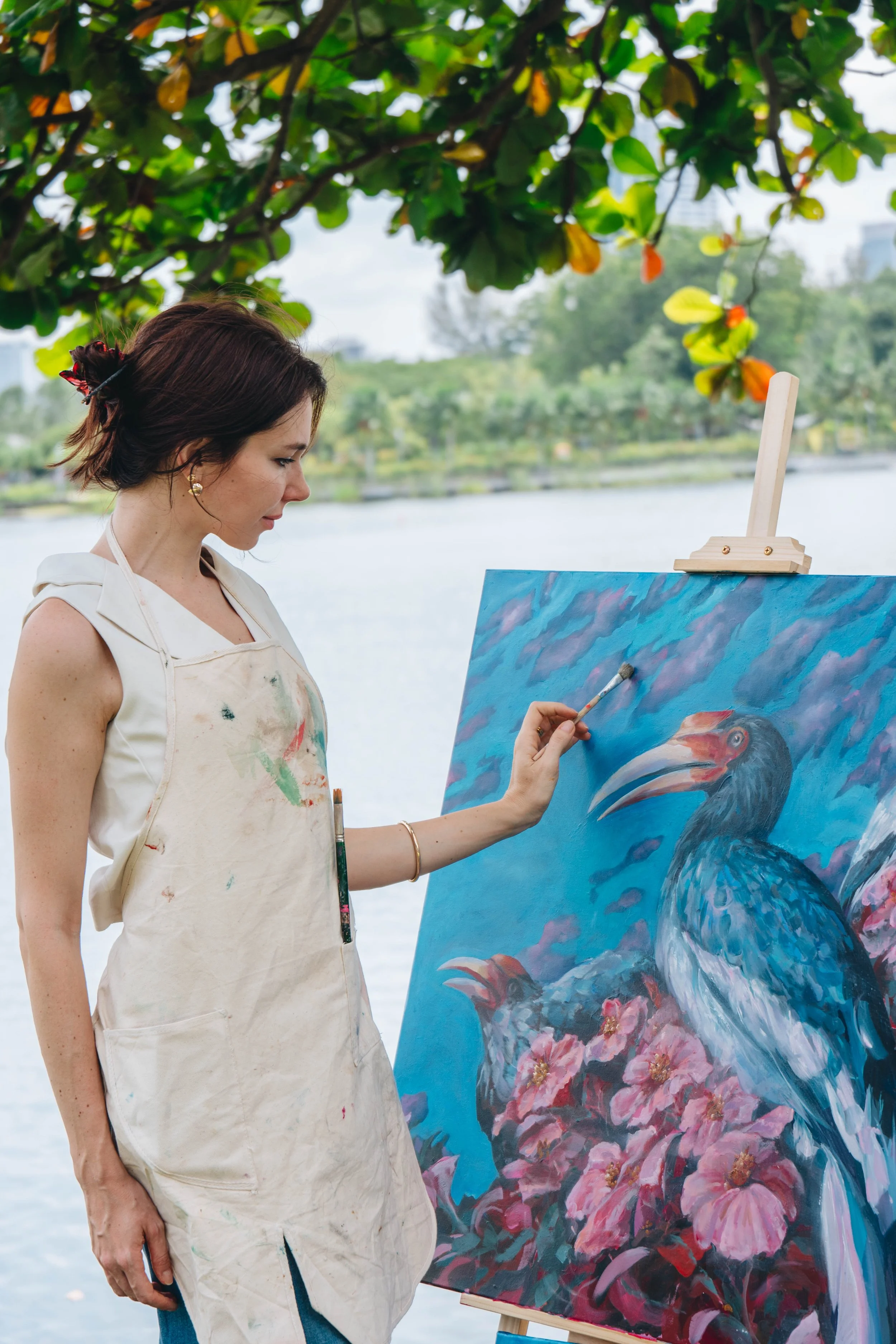 A woman painting a colorful picture of toucans and pink flowers on an canvas, outdoors near water with trees in the background.