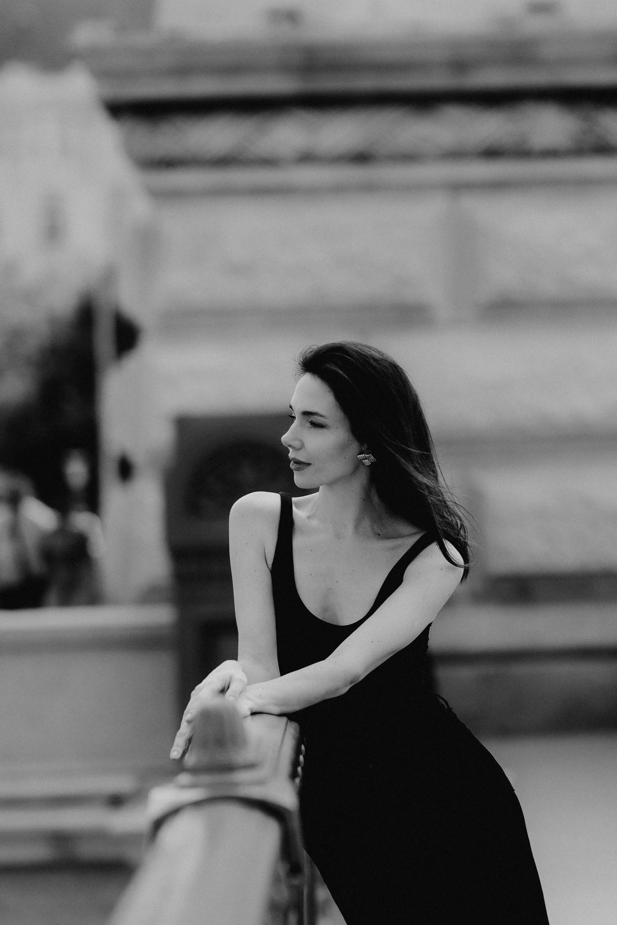 A black-and-white photo of a woman with dark hair looking to the right, wearing a sleeveless black dress, standing by a railing in an indoor setting.