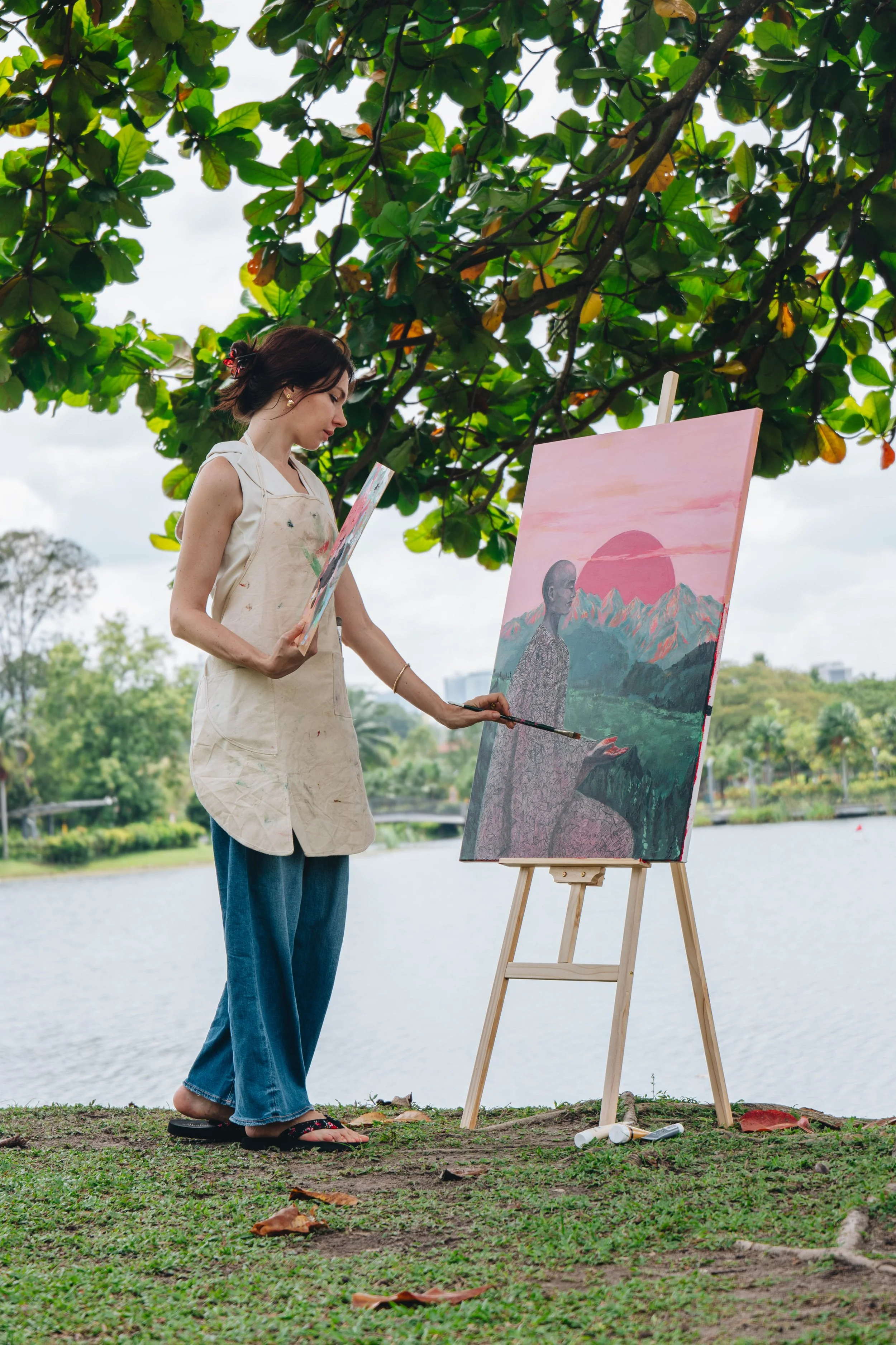 A woman painting outdoors beside a lake, with trees and mountains in the background, under a large leafy tree.