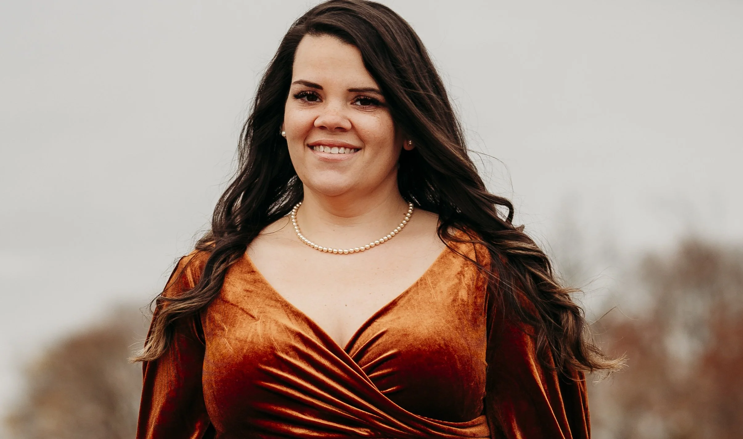 A woman with long dark hair, wearing a pearl necklace and an orange dress, smiling outdoors against a blurred background.