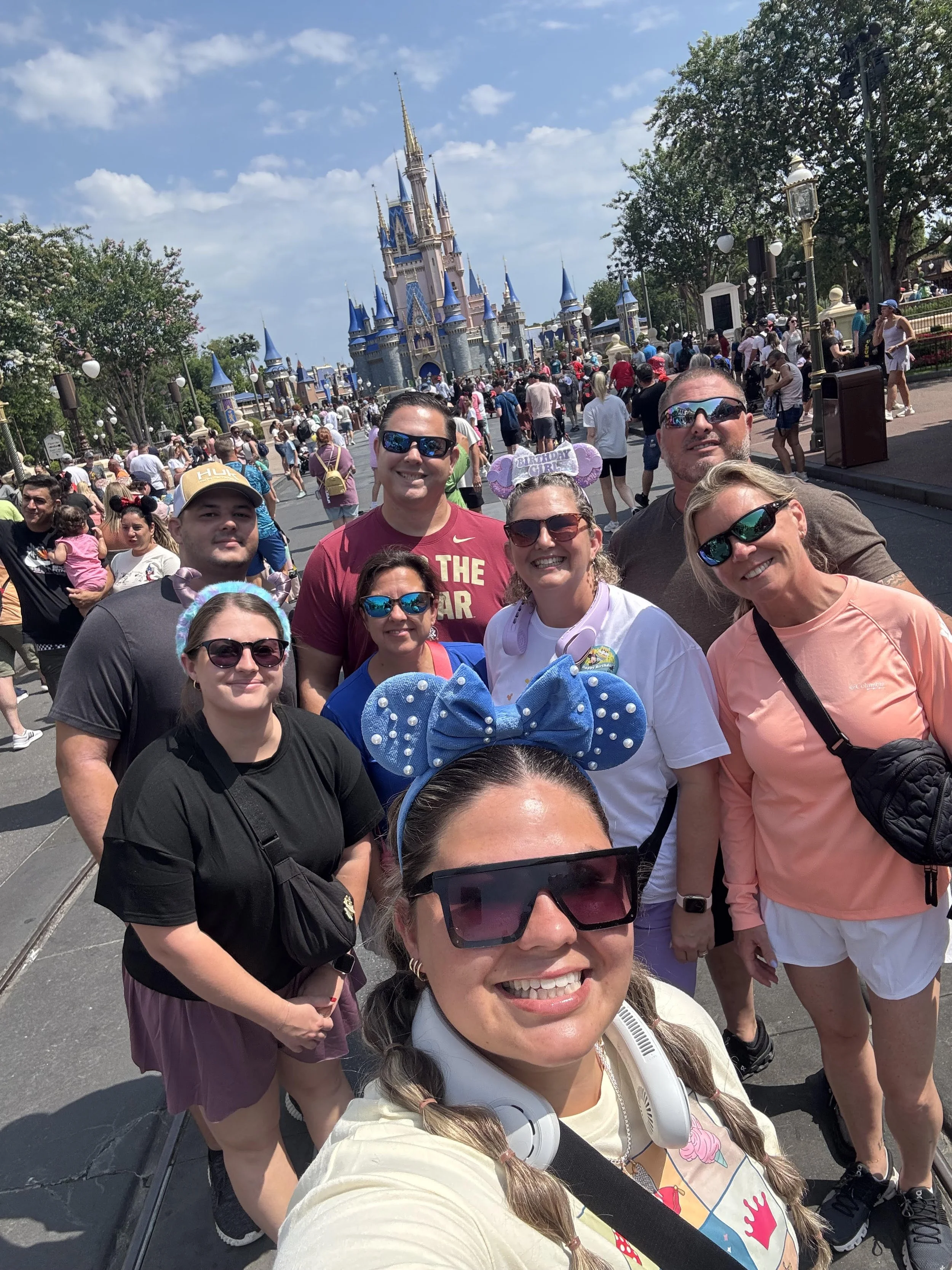 Group of people taking a selfie in front of Sleeping Beauty castle at Disneyland on a sunny day with a clear sky, surrounded by other visitors.