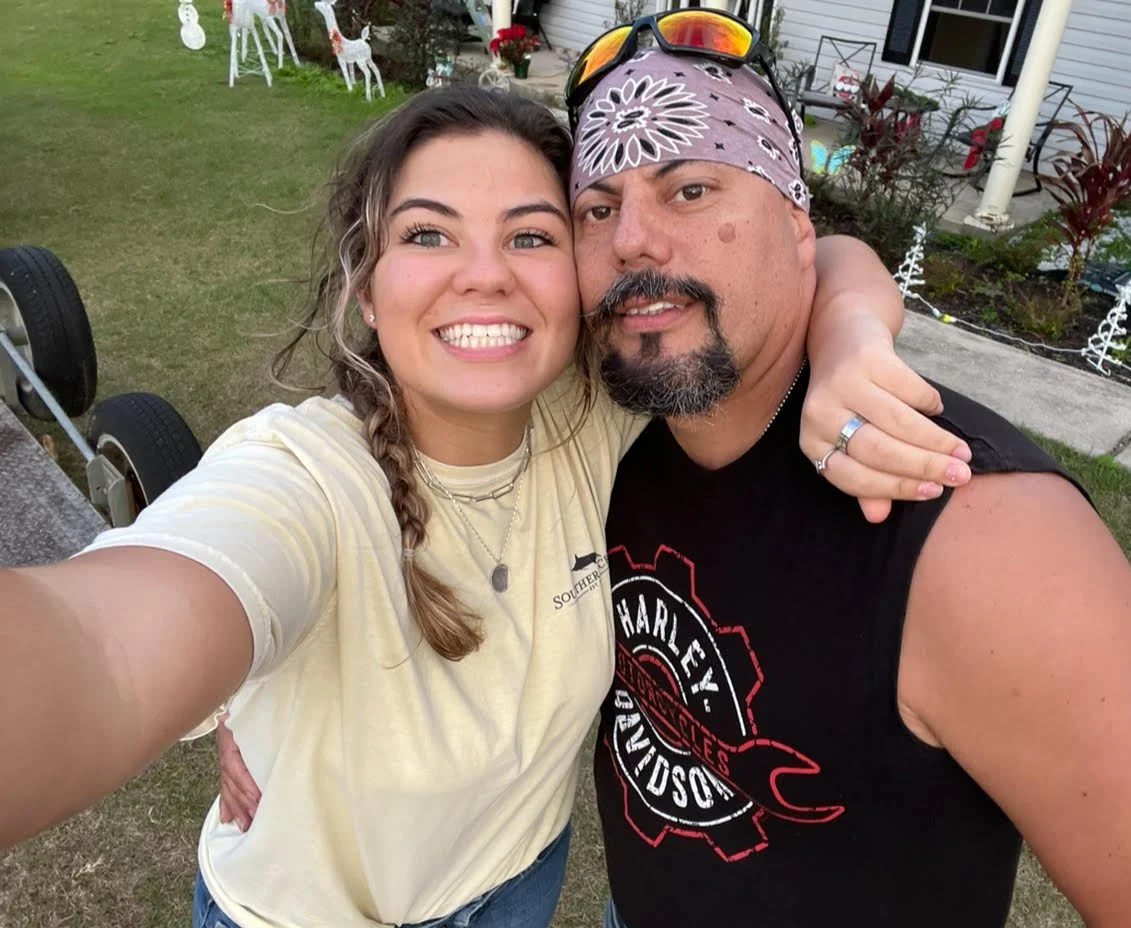 Two people taking a selfie outdoors, smiling and hugging each other. The woman has light brown hair in a braid, wearing a yellow T-shirt, and several necklaces. The man has a beard, wearing a bandana and sunglasses on his head, with a black sleeveless shirt that has a Harley Davidson logo. There are Christmas decorations in the background, indicating a festive setting.
