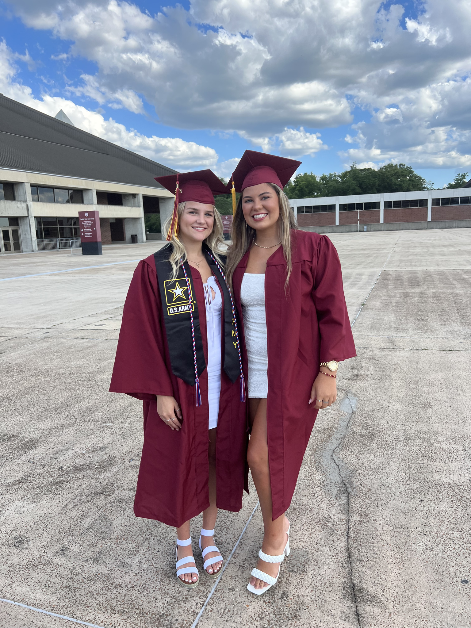 Two young women in maroon graduation gowns and caps standing outdoors on a concrete surface, smiling, with a partly cloudy sky and a building in the background.