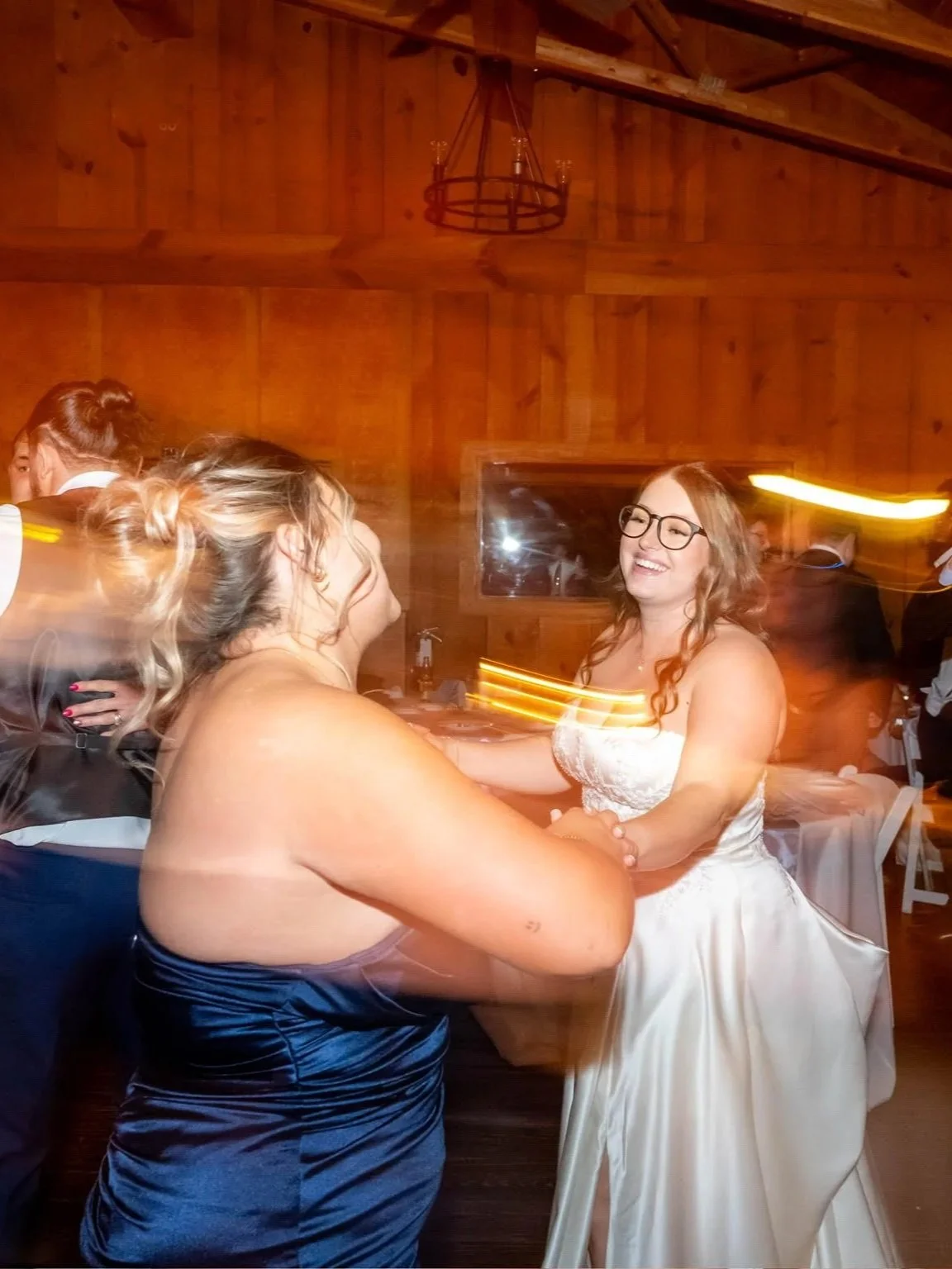 Two women dancing and smiling at a wedding reception, with a woman in a white wedding dress and glasses.