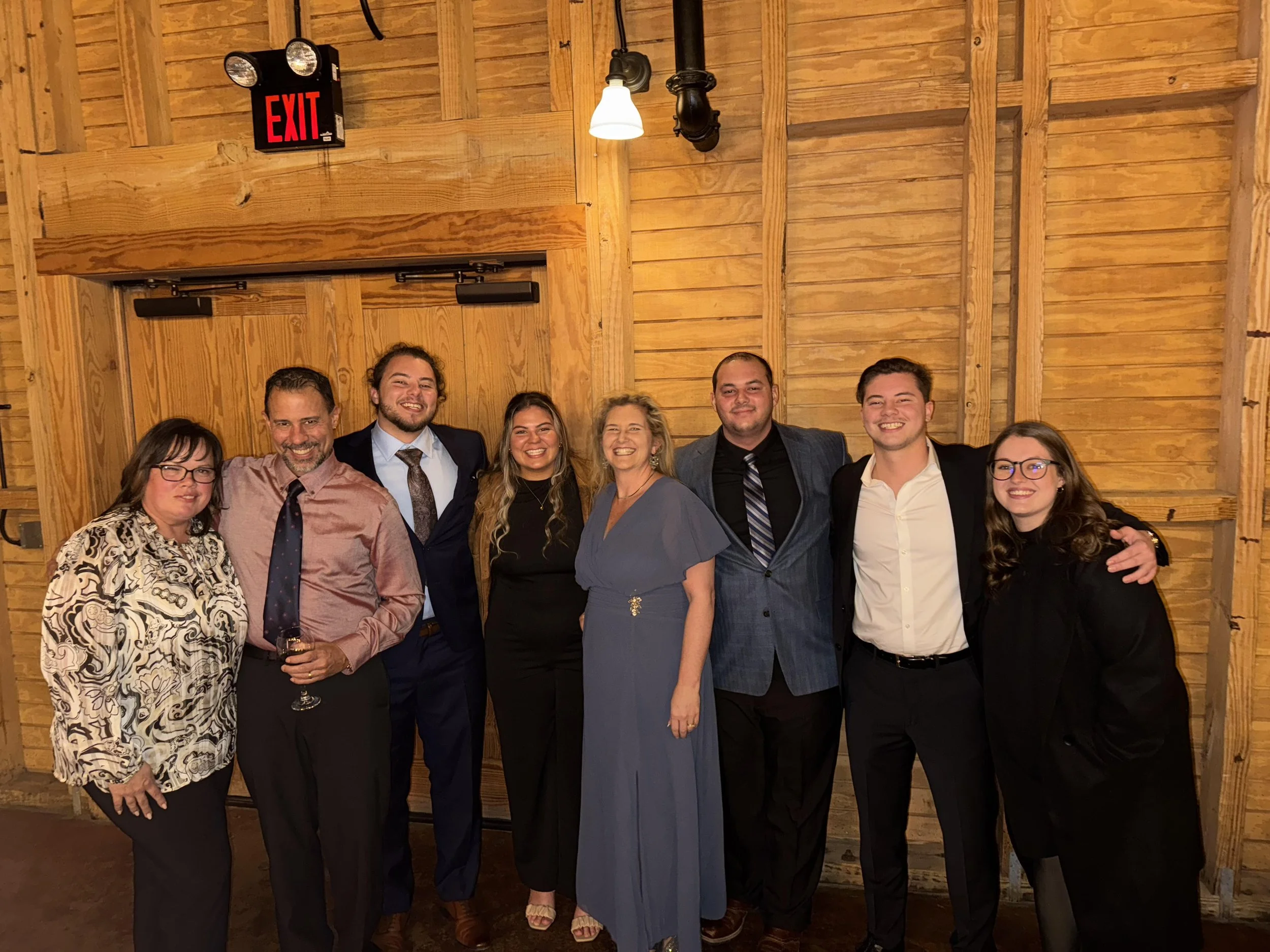 Group of eight people standing together inside a wooden room, smiling for the camera, with a red exit sign above a closed sliding door.