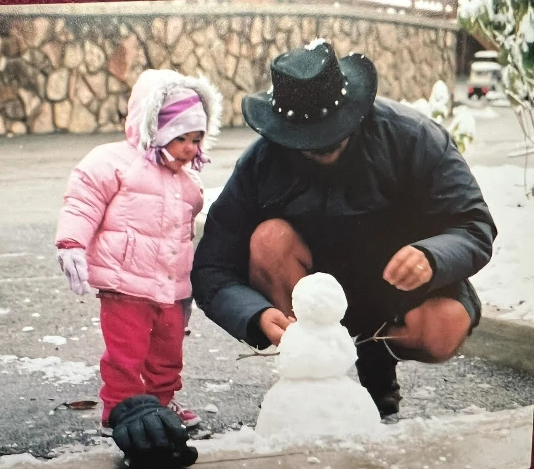 A man and a young girl building a small snowman outdoors in winter, with a stone wall in the background.