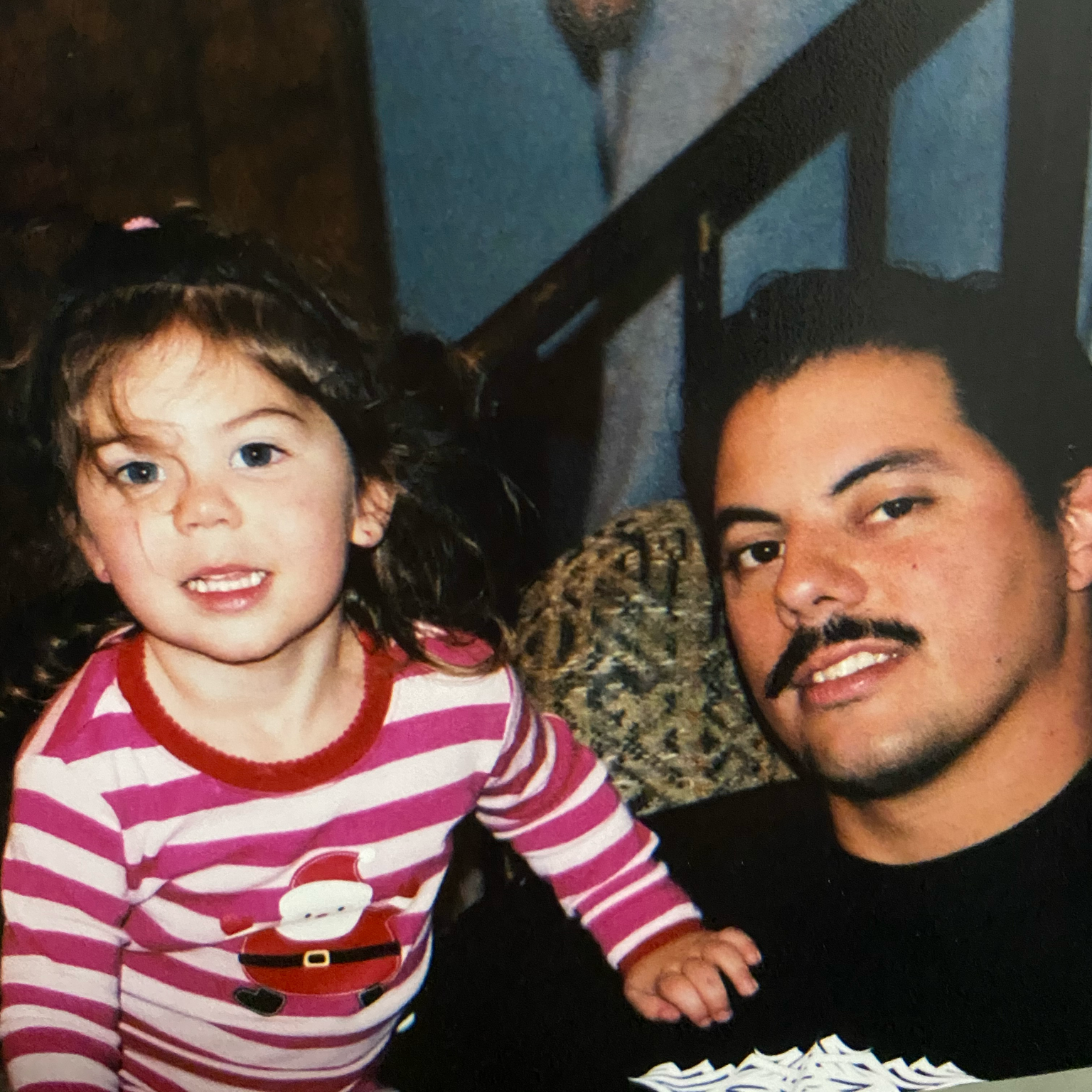 A young girl with curly hair and a man with dark hair and a mustache, both smiling, indoors near a staircase.