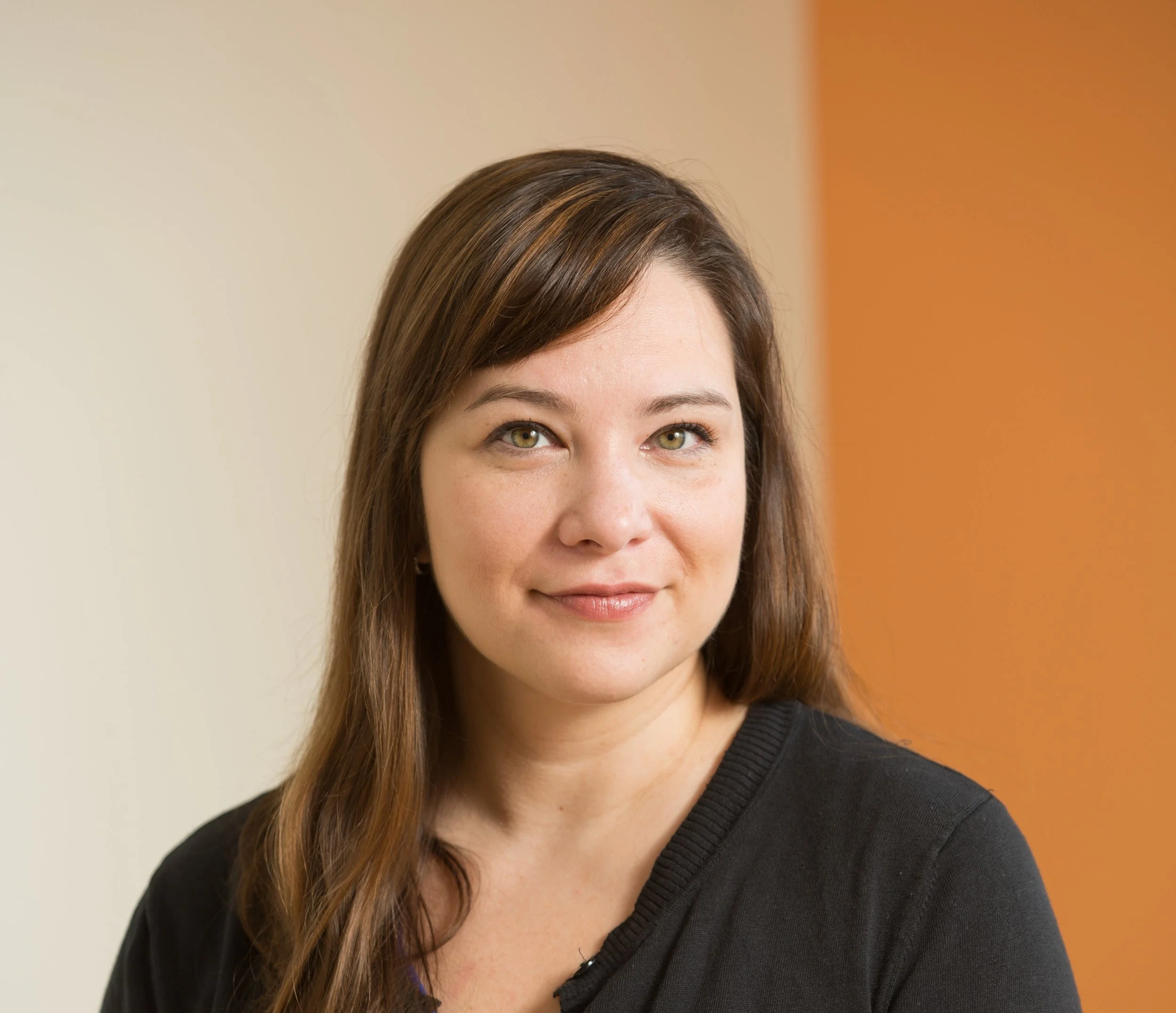 A woman with brown hair and hazel eyes, wearing a black top, smiling and looking slightly to her right, with a beige and orange background.