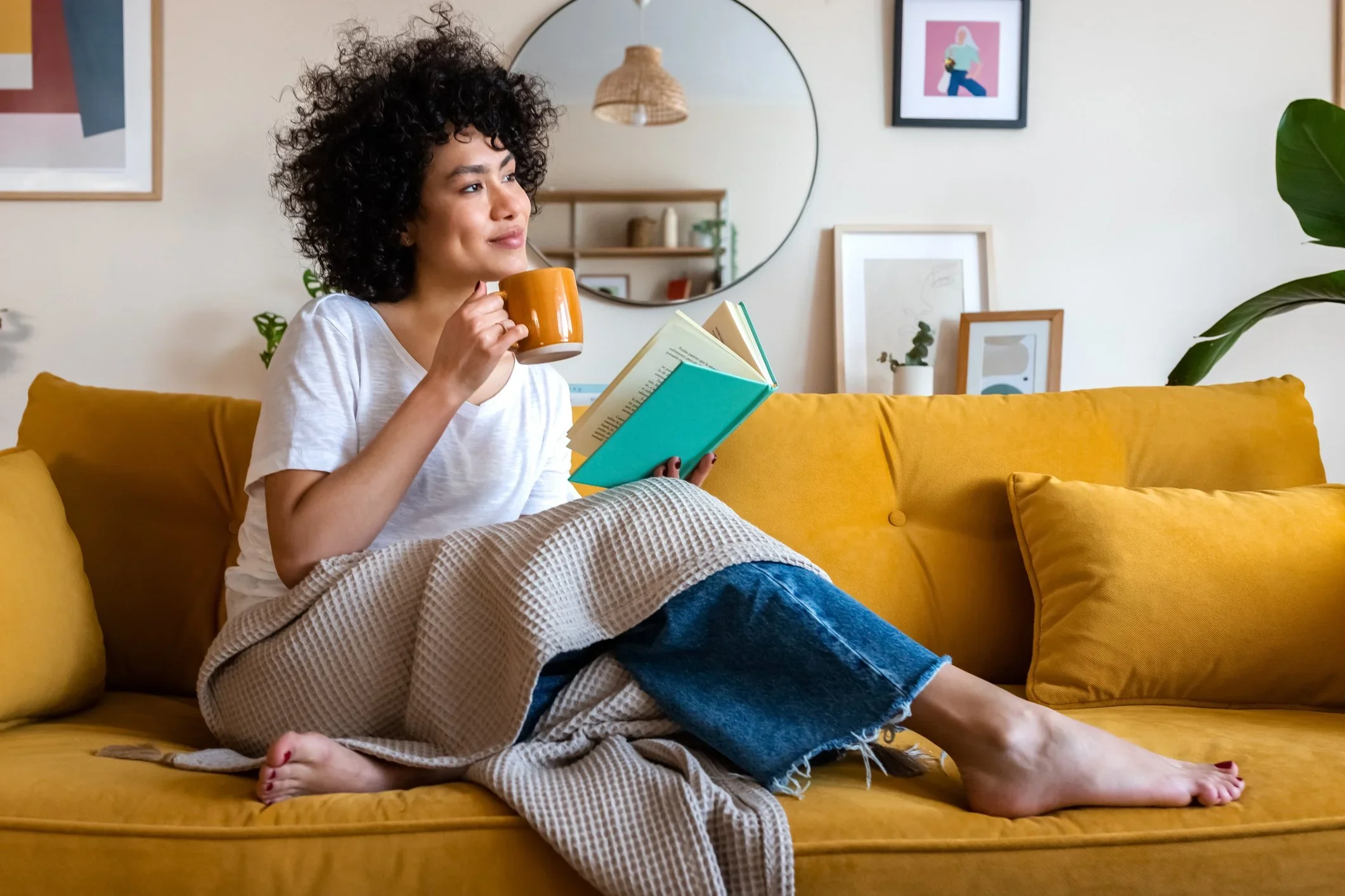 A woman with curly hair sitting on a yellow couch, holding a yellow mug in one hand and a book in the other, enjoying a relaxing moment in a cozy, decorated living room.