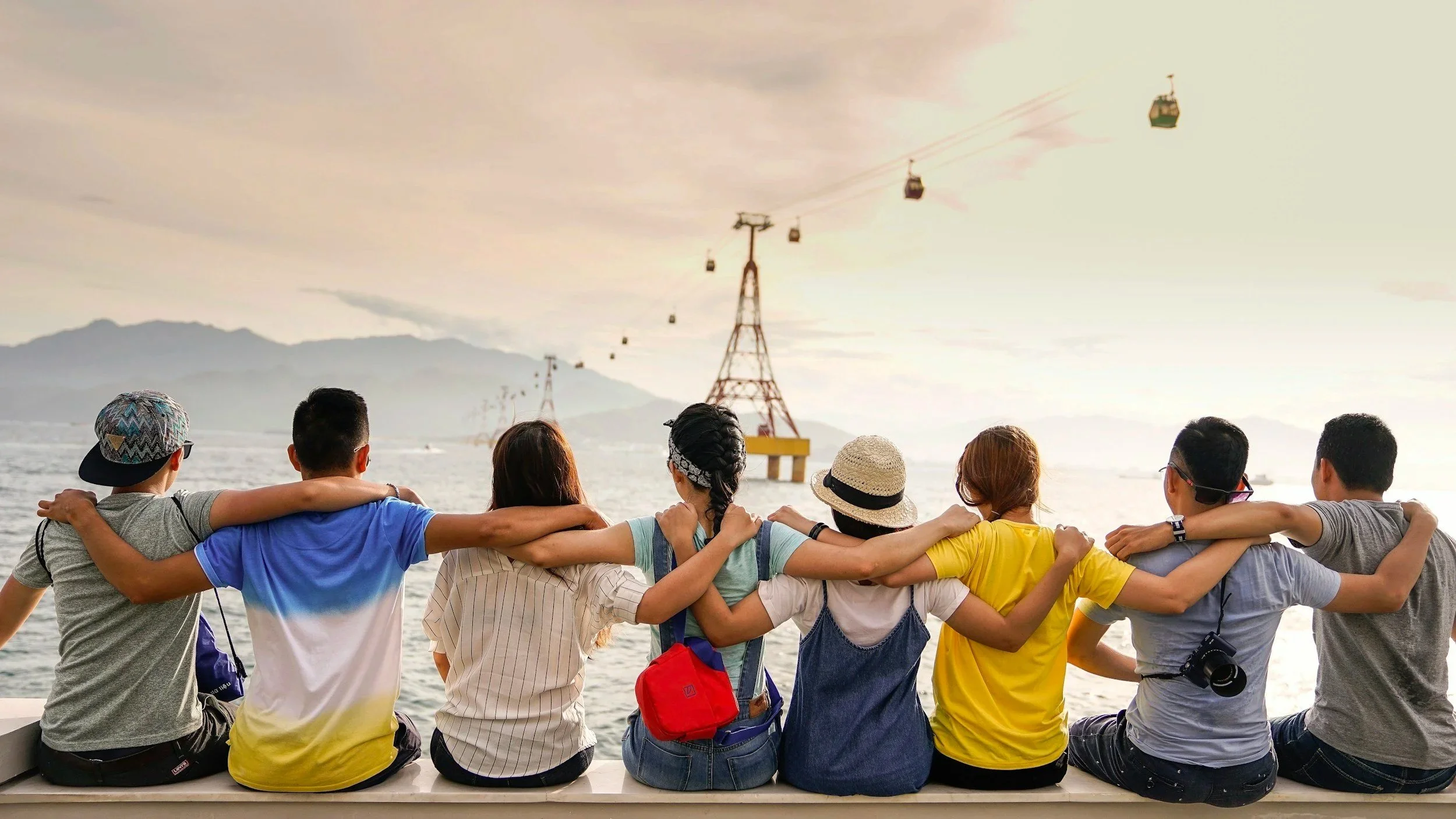 Group of friends sitting on a bench with arms around each other, watching cable cars in the water near mountains during sunset.