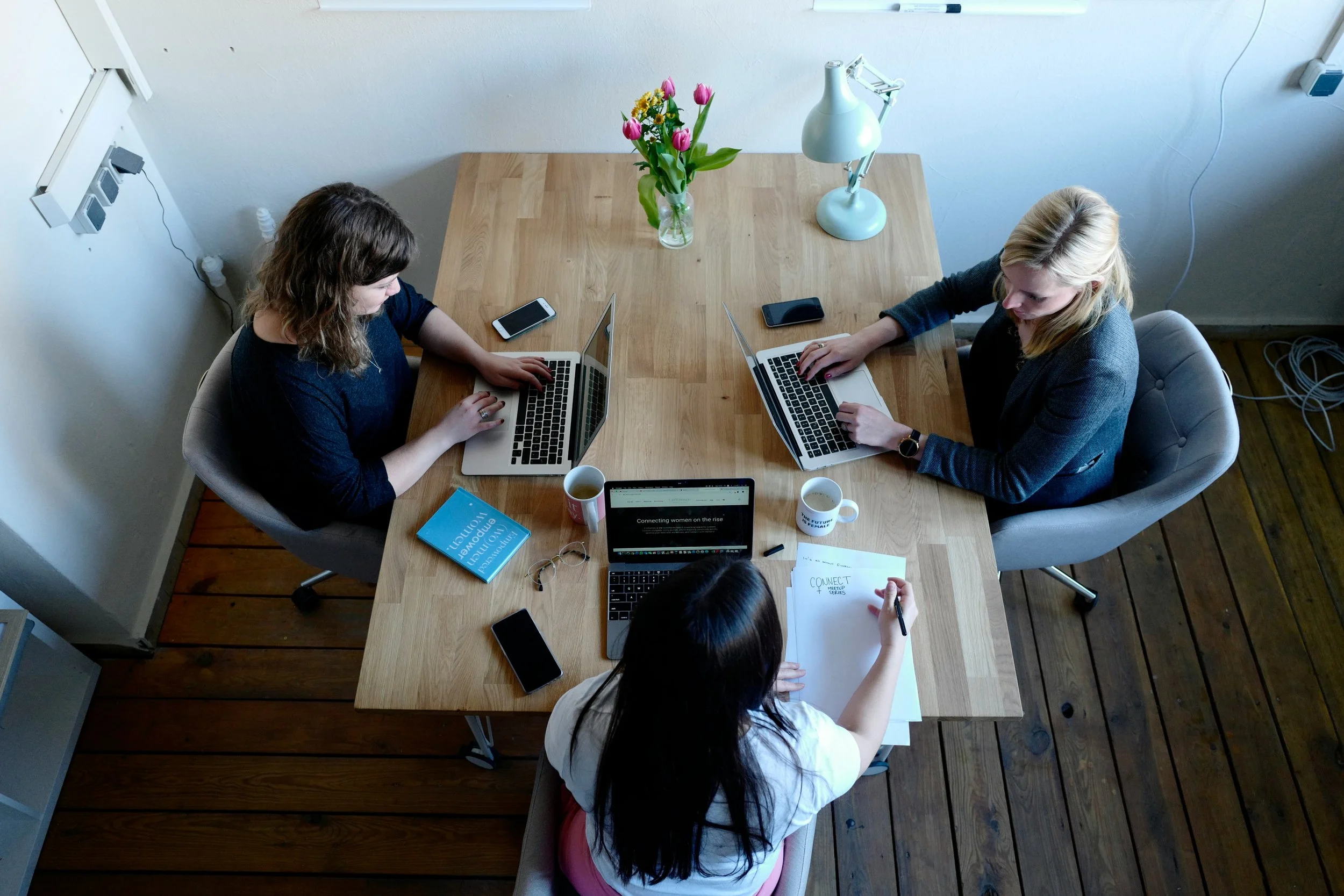 Three women working at a wooden conference table with laptops, notebooks, and coffee mugs in a well-lit room with wooden flooring and a vase of pink and purple flowers.