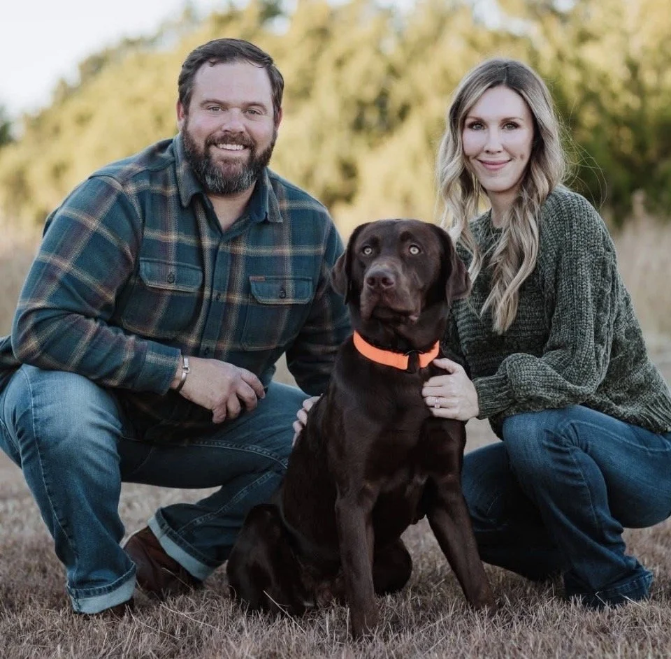 A man and woman kneeling outdoors with a chocolate Labrador retriever dog with an orange collar, on a grassy field with trees in the background.