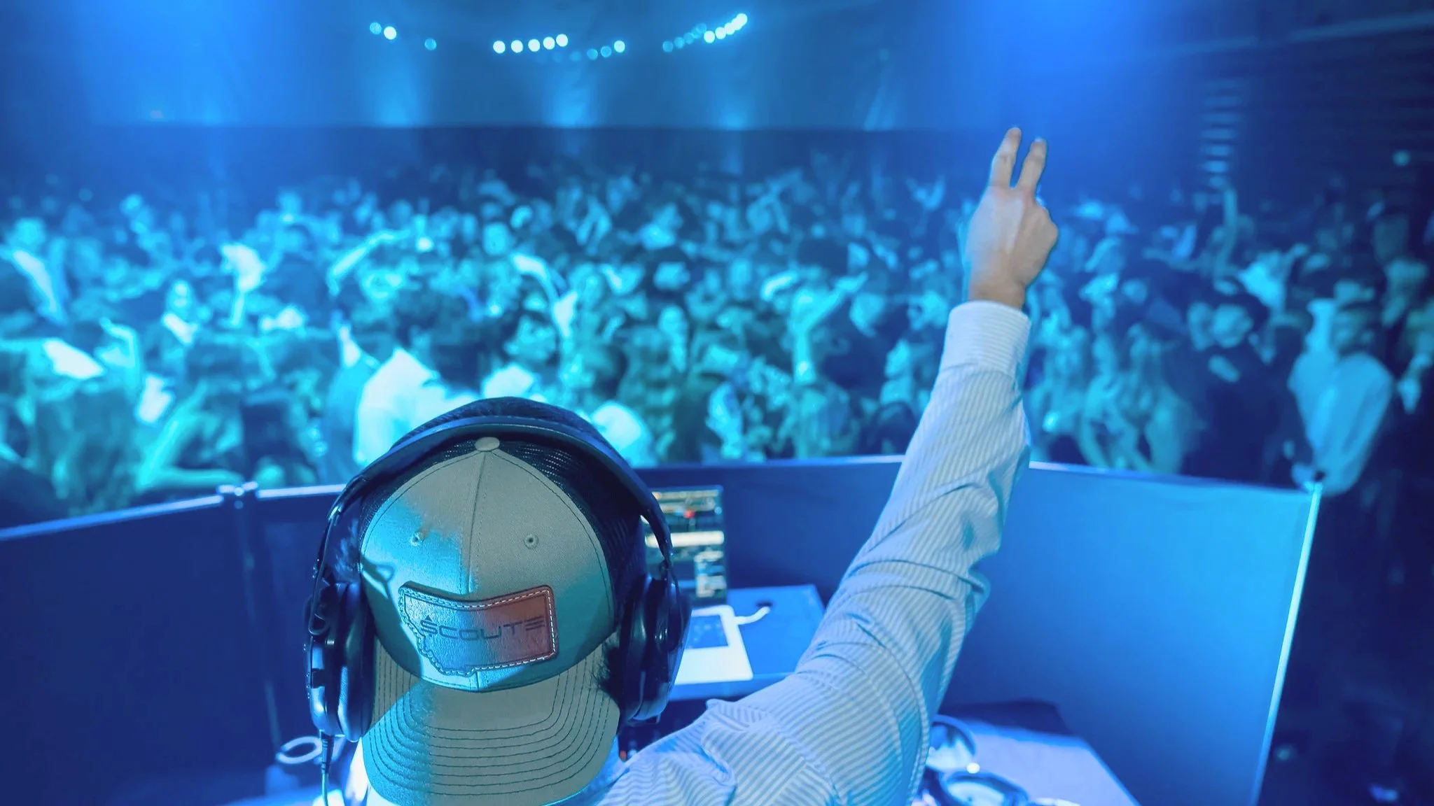 School dance DJ at the DJ booth with headphones, raising his hand, in front of a large crowd at a school dance. The DJ is energizing the crowd.