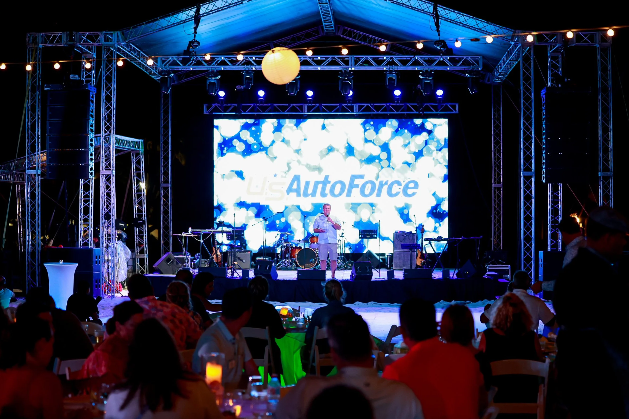 A stage with a large screen displaying 'U.S. AutoForce' in blue text, set up with musical instruments and surrounded by metal trusses. An audience is seated at tables with drinks, watching the performance at night.