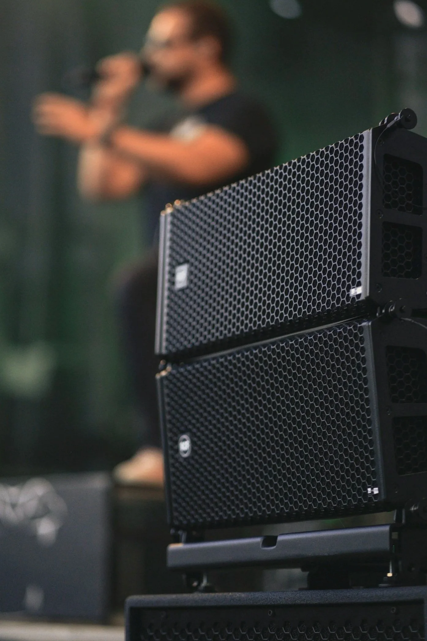 A large black speaker with a honeycomb grille pattern in the foreground, and a blurry person with glasses in a dark shirt speaking into a microphone in the background.