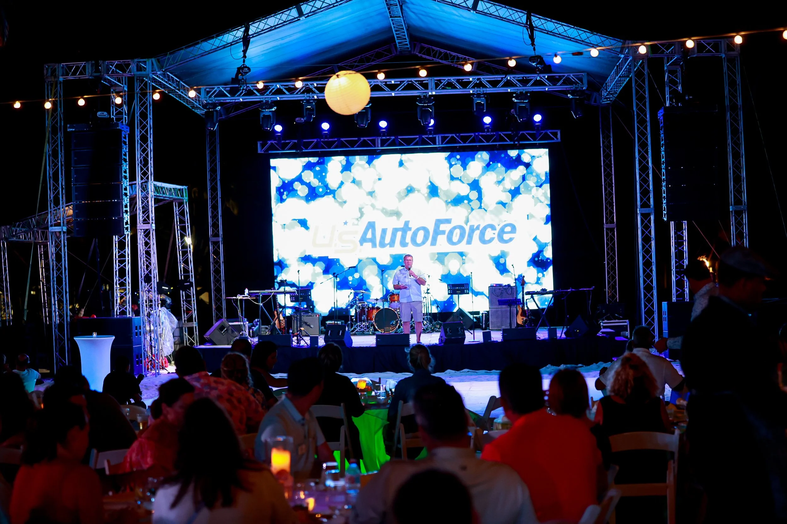 Scene from an outdoor corporate event with a stage and an audience. The stage is lit with blue lighting, has a large LED screen displaying the company logo, and musical equipment. People are seated at tables with candles, watching the performance.