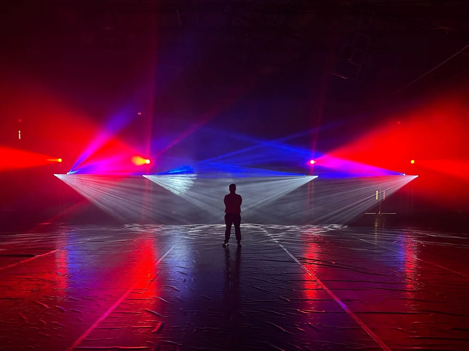 A person standing in a school gymnasium with colorful stage lights casting red, blue, and white beams, decorated for a large school dance.