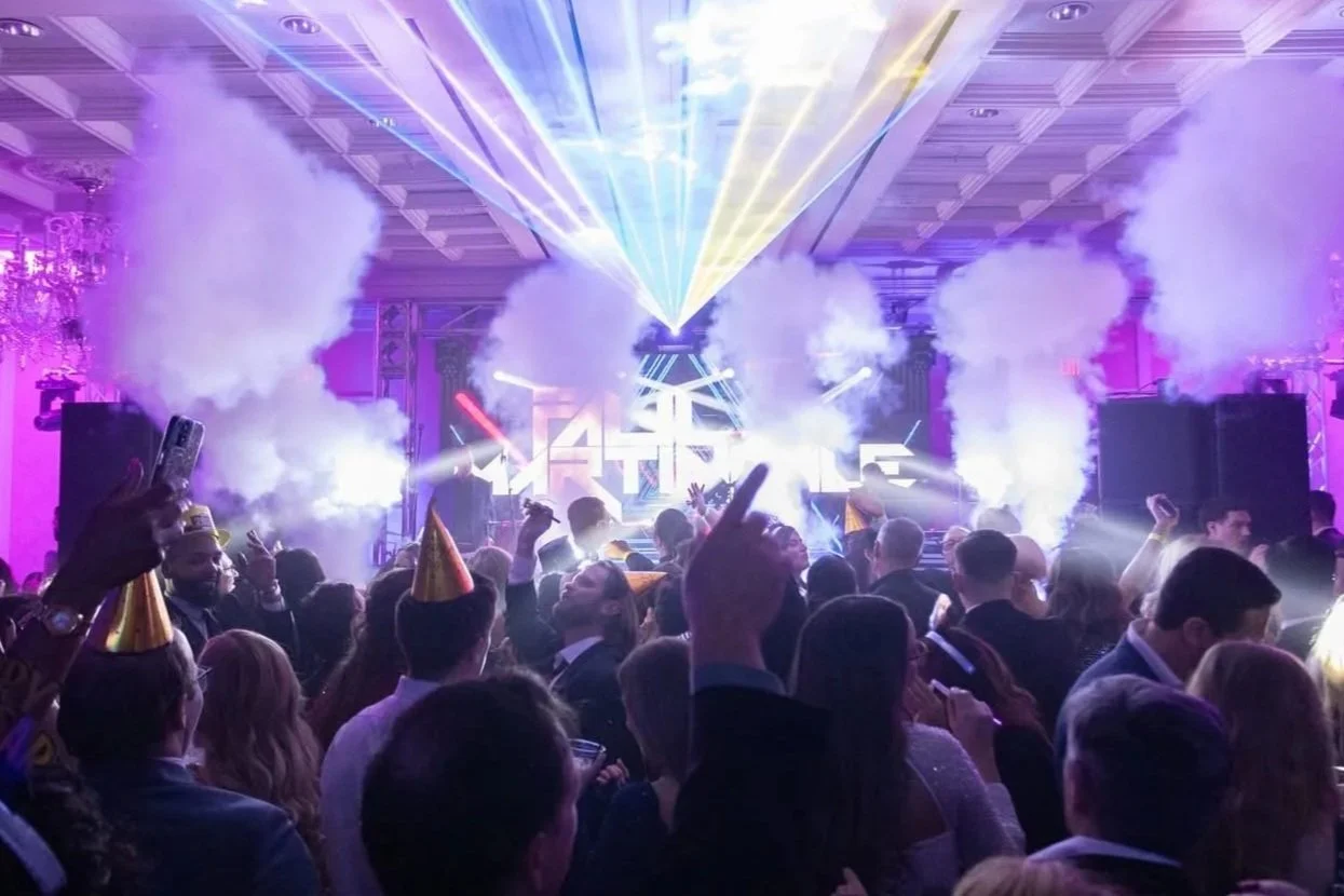 Crowd of people at a celebration event with colorful laser lights, smoke effects, and some wearing party hats, dancing and enjoying the music.