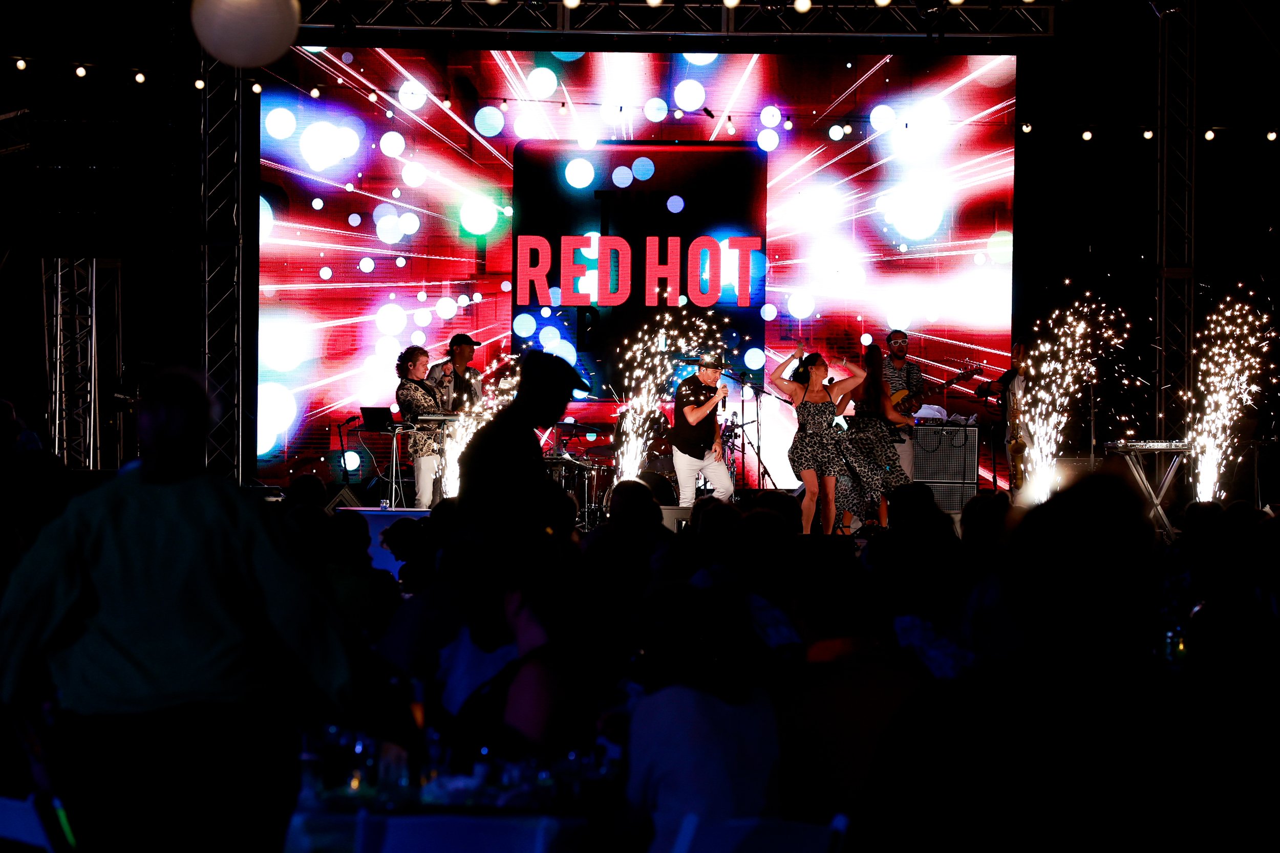 A live performance with a band and dancers on stage, illuminated by bright lights and fireworks, with a large screen in the background displaying the words "RED HOT."