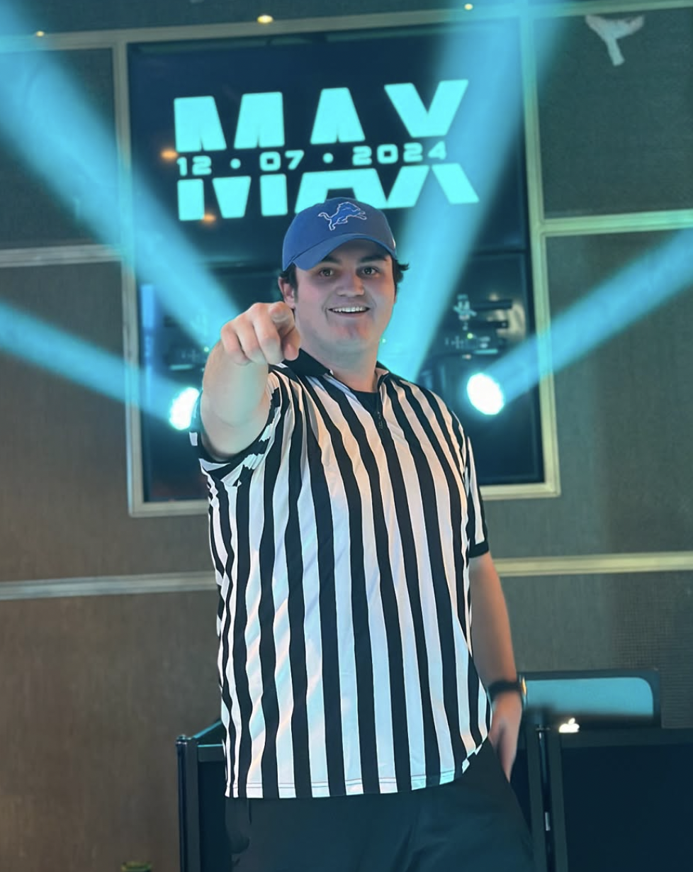 A smiling man points toward the camera, standing in a room with blue and teal lighting.