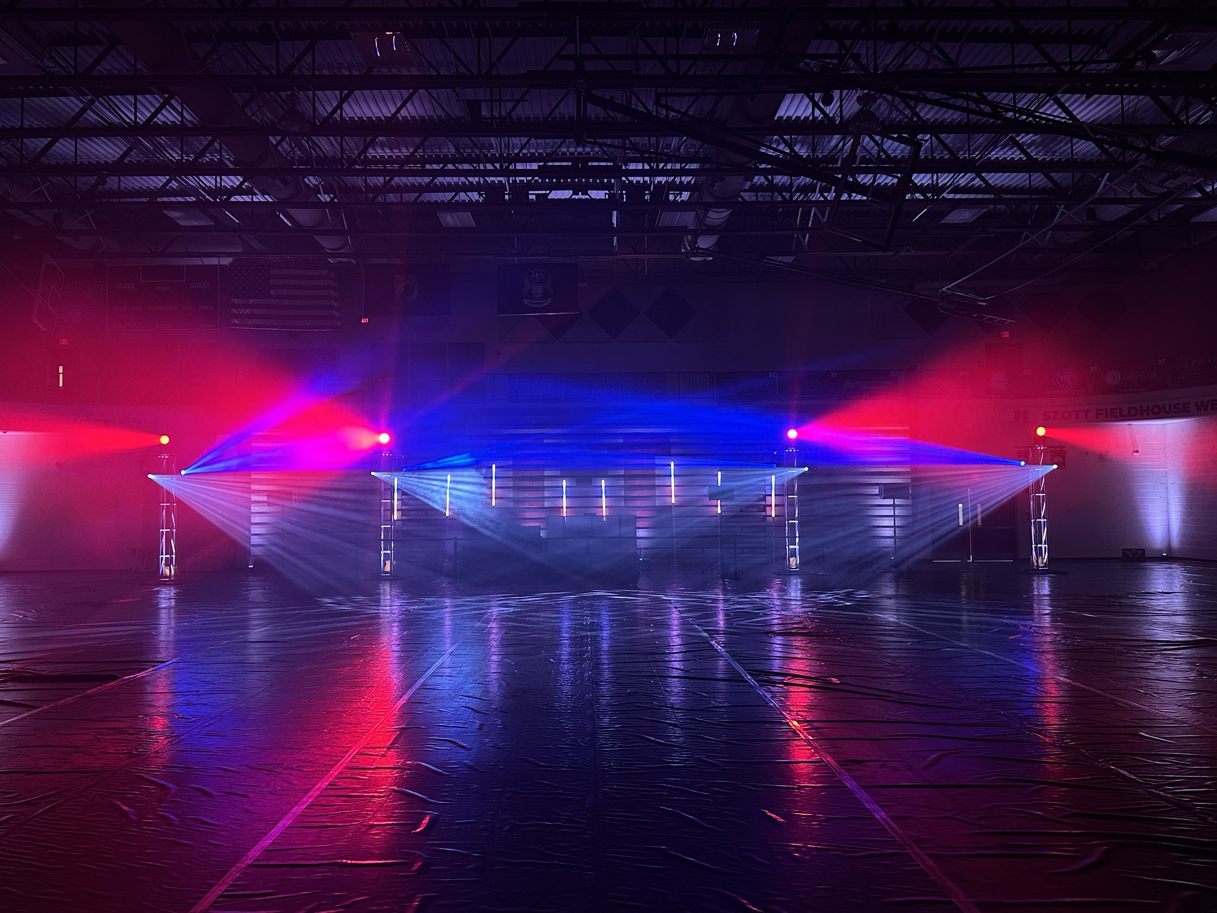 An school dance stage setup with vibrant red, pink, and blue lighting effects, reflecting off a glossy floor, with a visible ceiling structure and banners in the background.