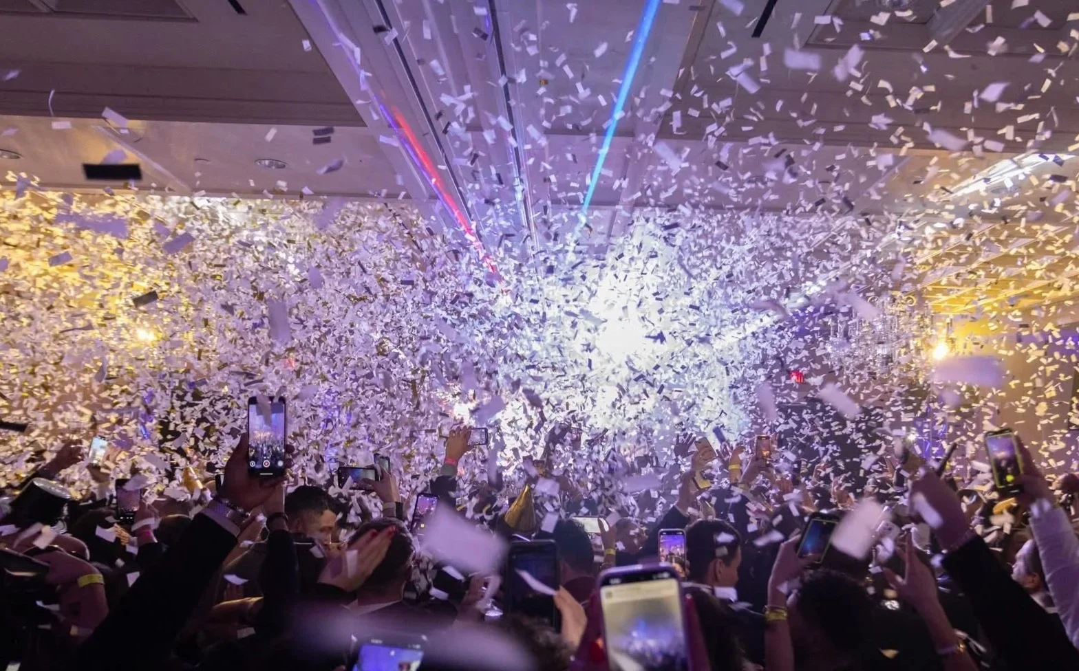 Crowd at an indoor event celebrating with confetti and party hats, many people capturing the moment on smartphones.