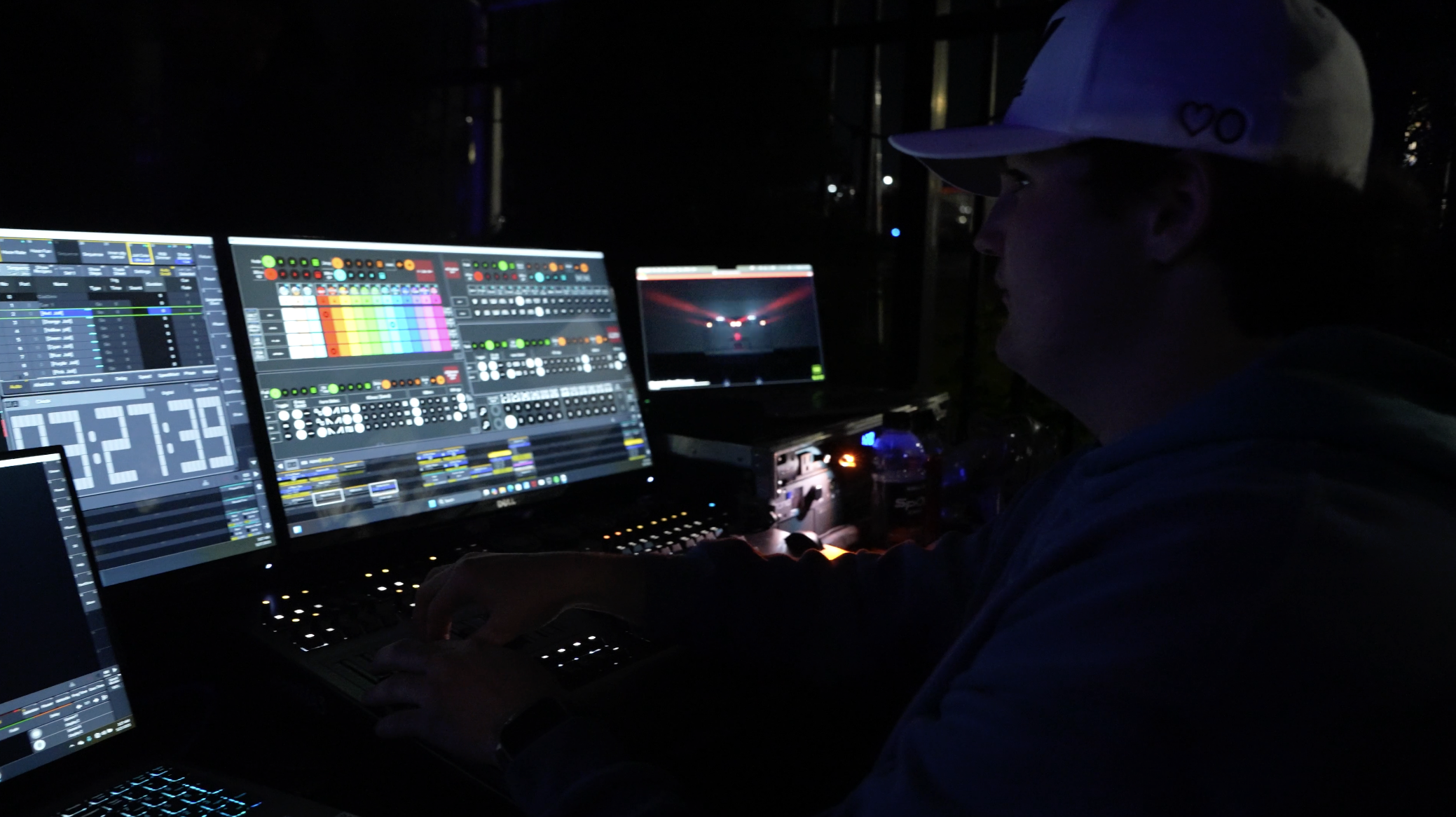 A person operating multiple computer screens displaying DJ or music production software with colorful waveforms and controls, in a dark environment, possibly during a performance or live event.