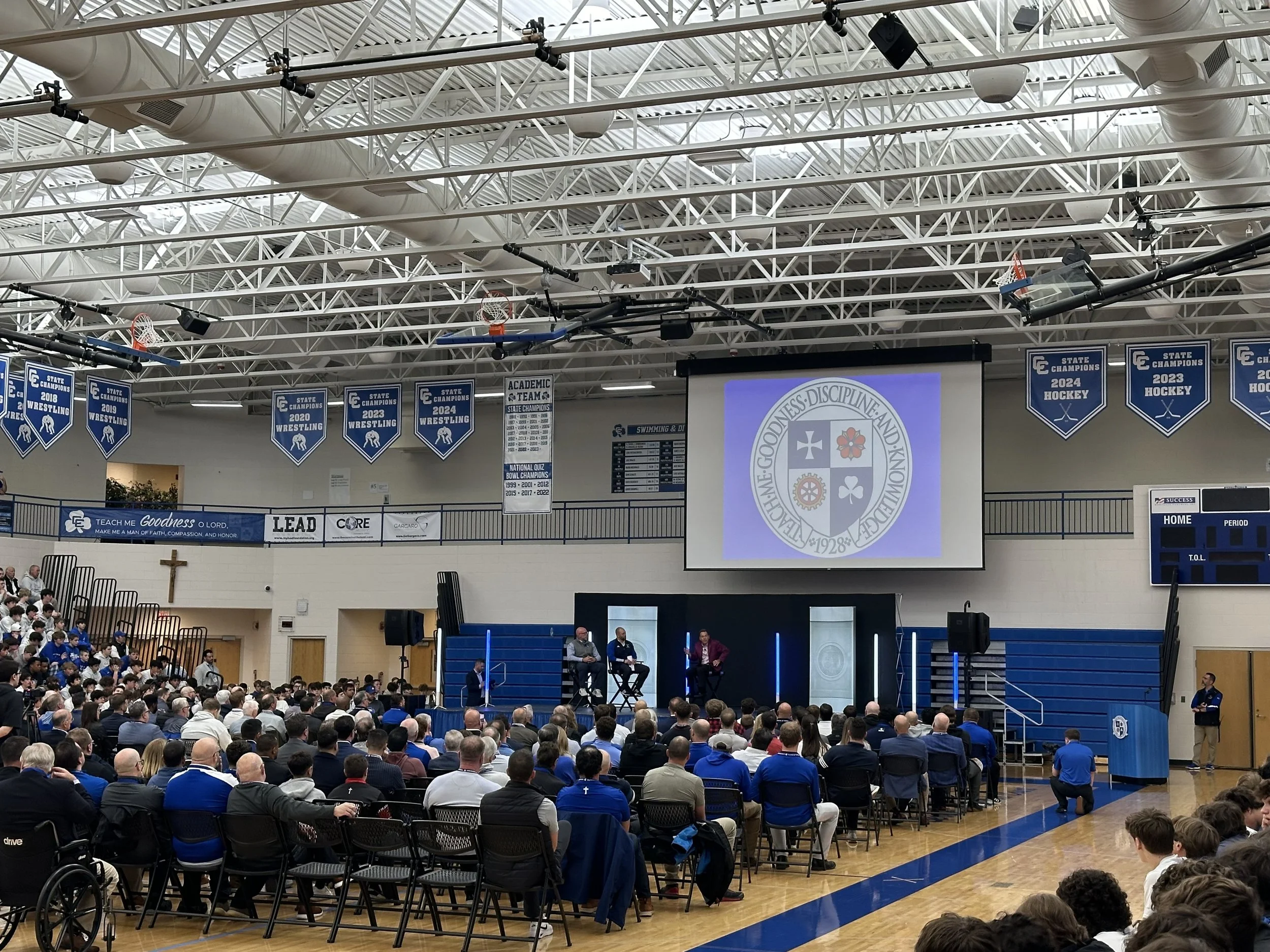 A large indoor gymnasium filled with people attending a presentation or panel discussion. There is a stage with two people seated and one person standing next to a large screen displaying a crest with religious symbols. The gym has banners hanging from the ceiling celebrating state championships in wrestling and hockey, and a cross is mounted on the wall.