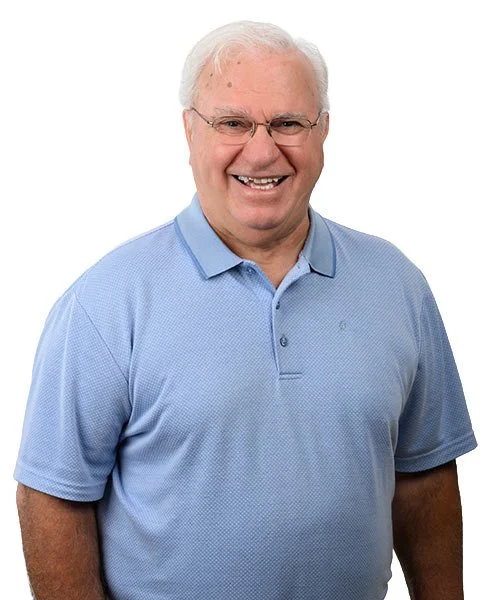 Un homme âgé souriant, portant une chemise bleue, contre un fond blanc.