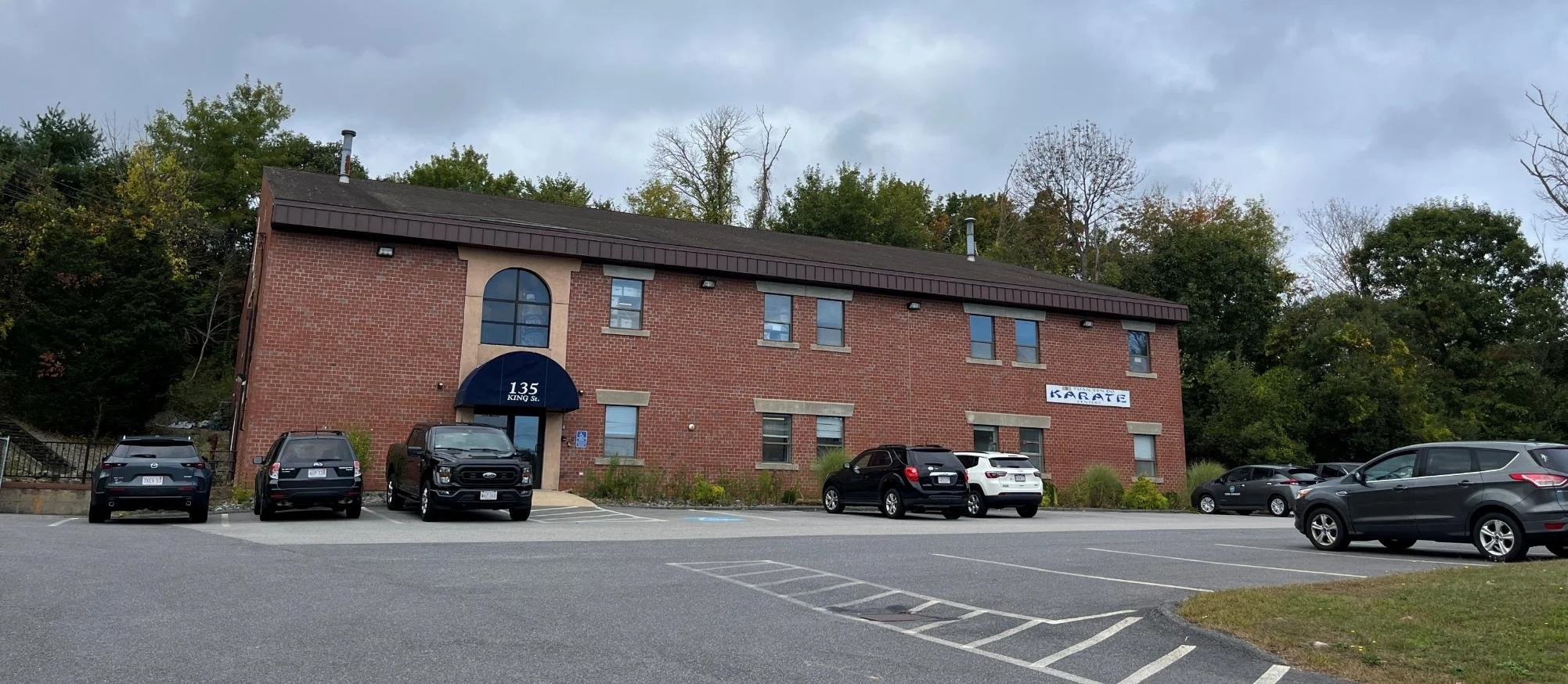 A brick building with a sign that reads '135 King St.' and a sign on the side that says 'KARATE'. There is a parking lot in front with several parked cars and a wooded area behind the building under a cloudy sky.