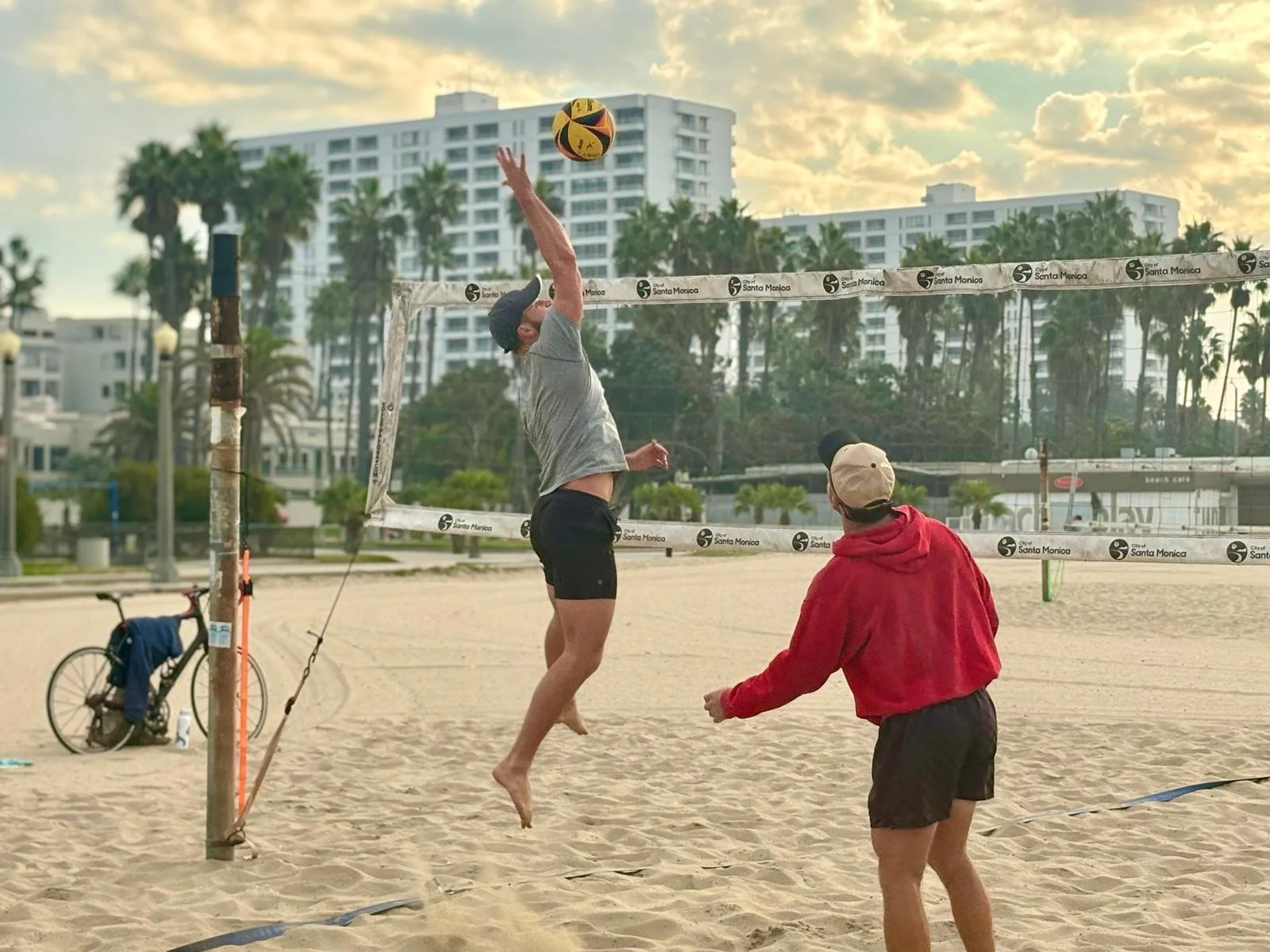 A person in a gray shirt and black shorts jumping to hit a volleyball at the beach volleyball court with coach in red hoodie instructing next to him.