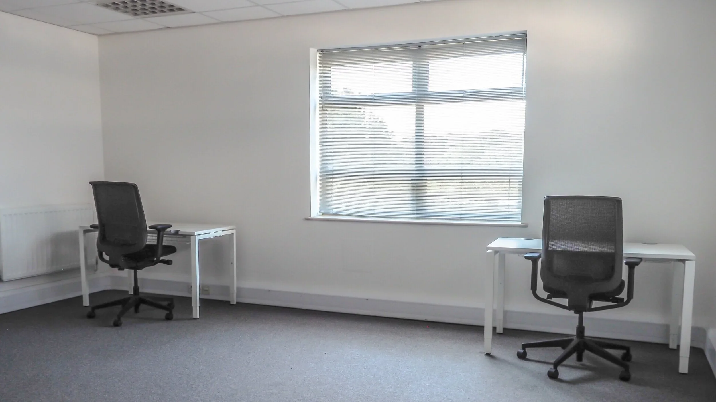 An empty office room with two white desks, each with a black office chair, and a window with closed blinds letting in daylight.
