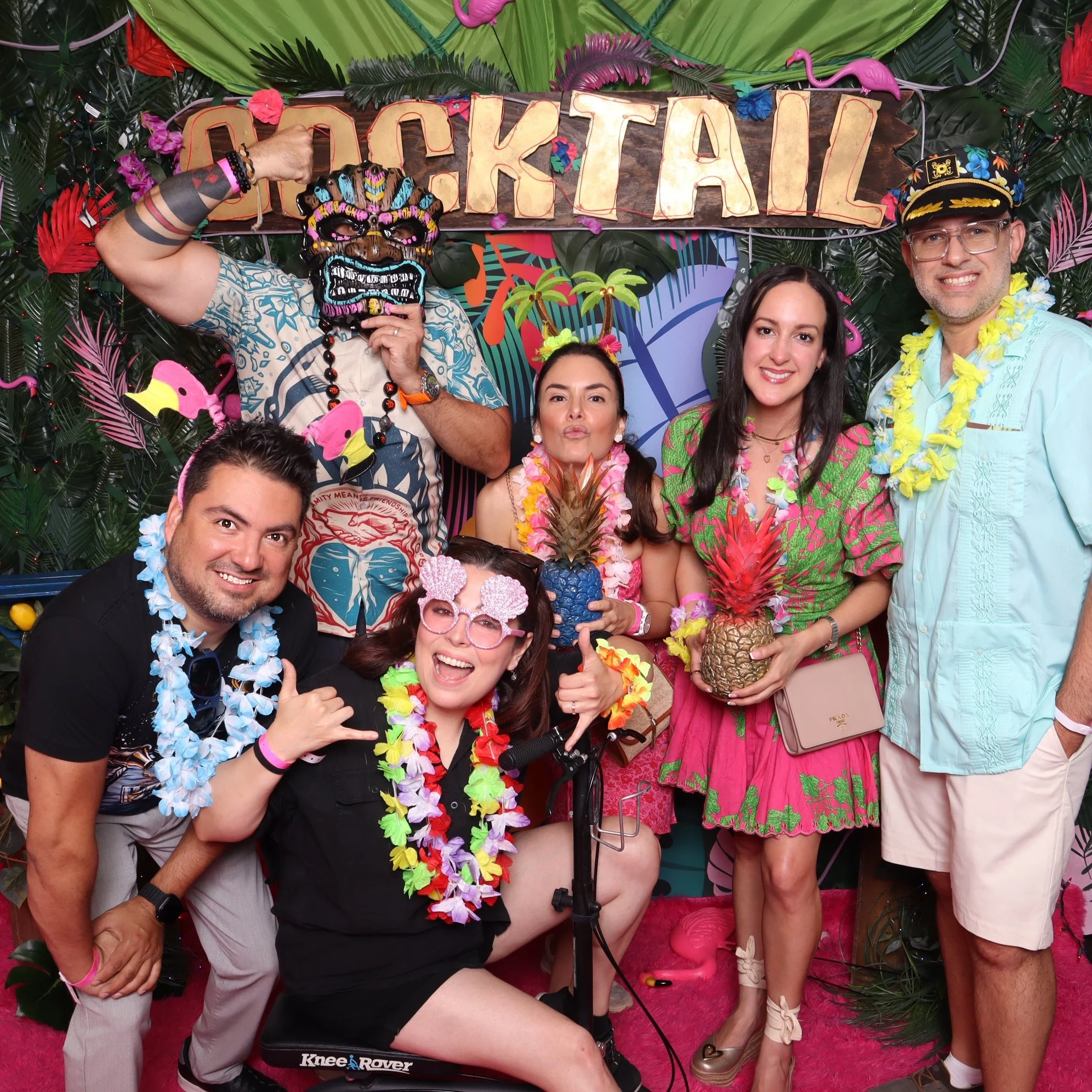 A group of six people in tropical-themed attire celebrating at a party with a sign that says 'Cocktail' and decorations of leis, pineapples, and flamingos.