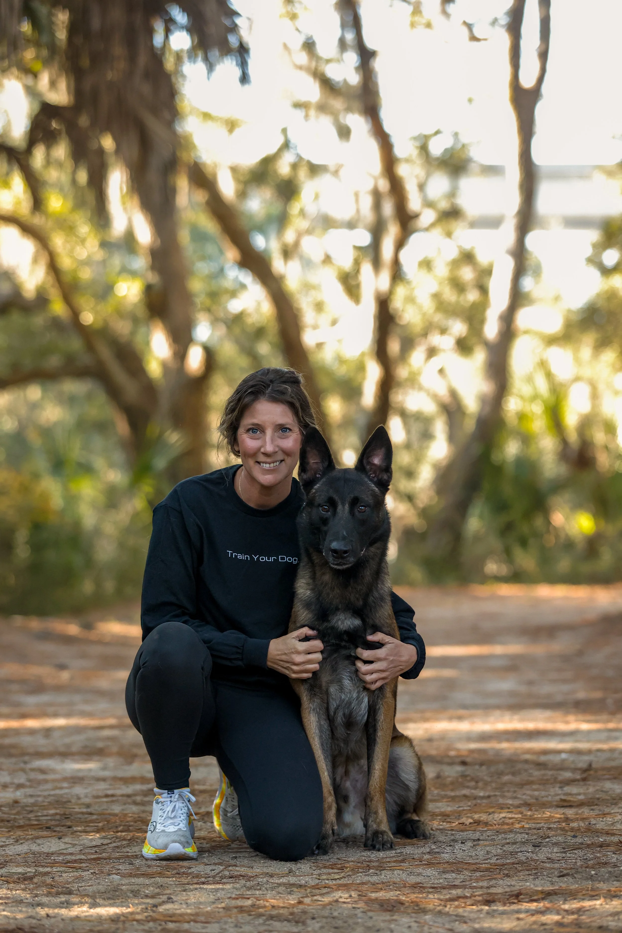 A woman kneeling on the ground in a wooded area, smiling while hugging a large German Shepherd dog sitting beside her.
