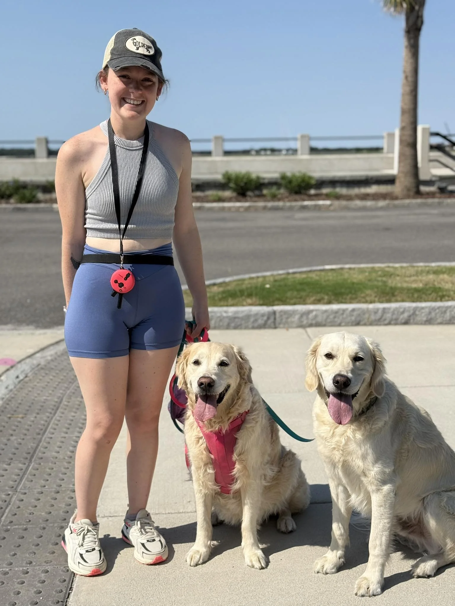 A smiling young woman in athletic attire standing outdoors with two golden retriever dogs, both sitting on the sidewalk with their tongues out, in a sunny environment near a street, with palm trees and a barrier in the background.
