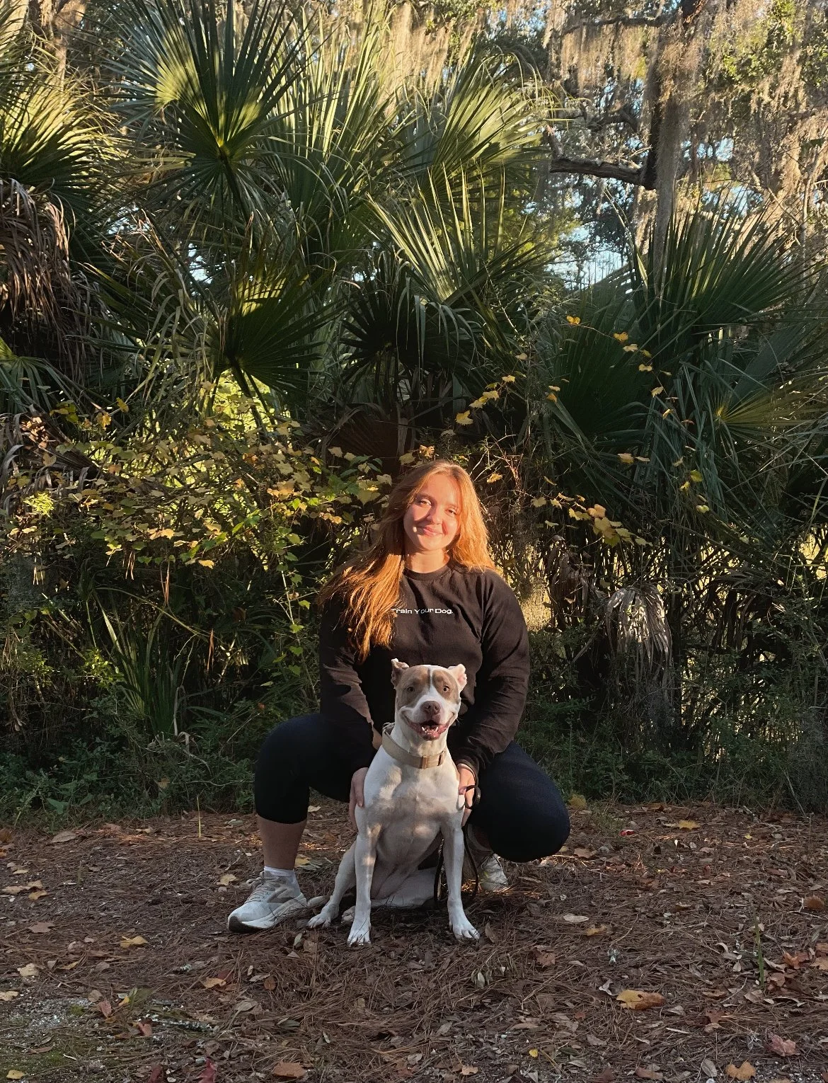 A woman in black athletic clothes squats on a forest trail, holding a white dog with a tan patch over one eye and a collar, in front of lush green foliage and trees.