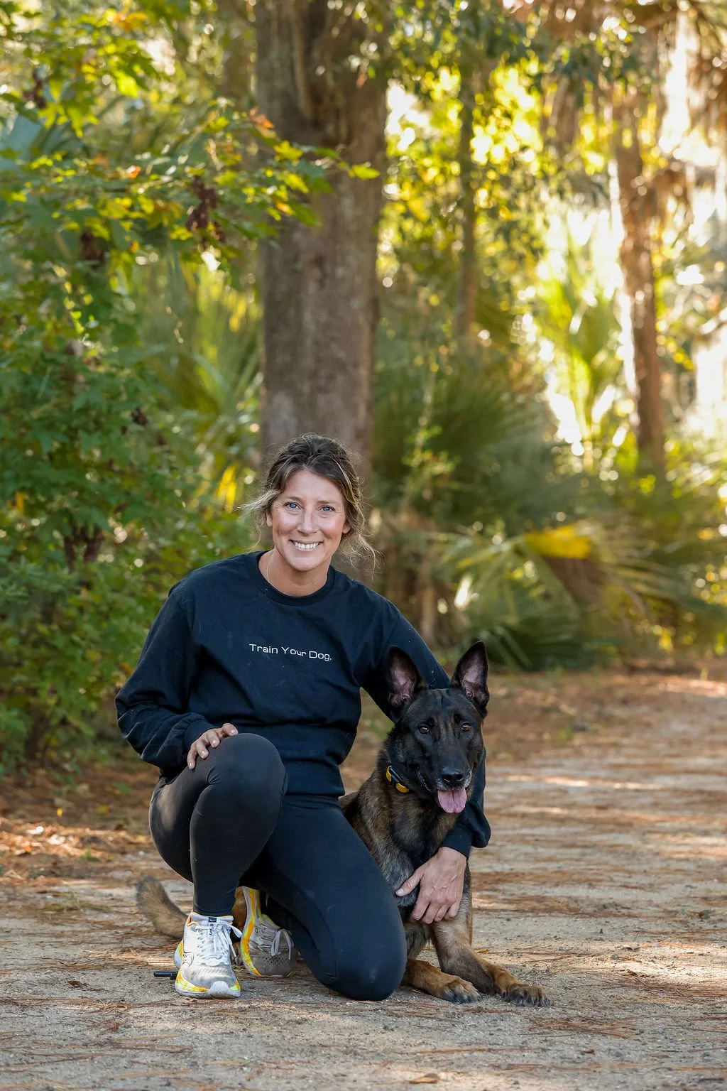 A woman crouching on a dirt trail in a forest with a large black and tan dog. The woman is smiling and wearing a black sweatshirt that says "Train Your Dog." The dog is sitting with its tongue out, next to the woman. Sunlight filters through the trees in the background.