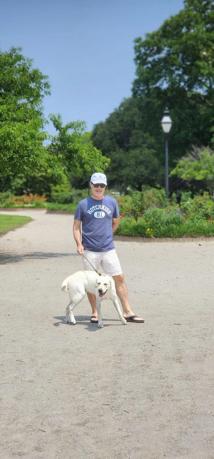 A man wearing sunglasses, a blue t-shirt with 'Hutchris' printed on it, white shorts, and flip-flops, walking a white dog with a happy expression on a sandy path in a park with green trees and a cloudy sky in the background.