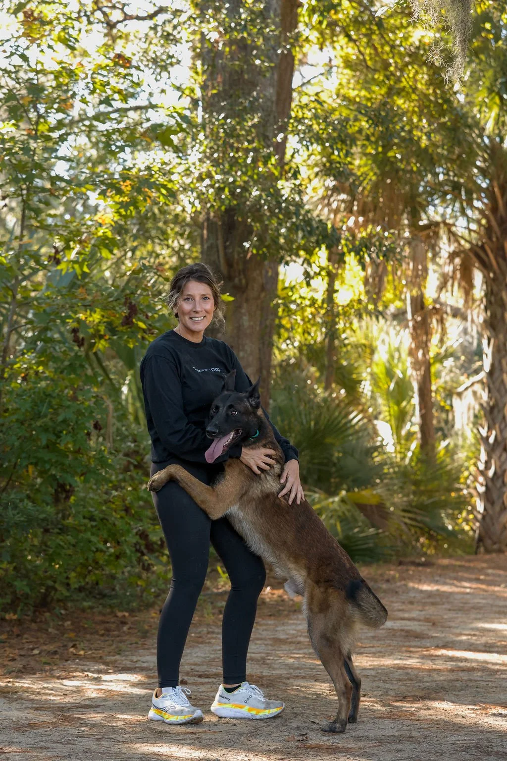 A woman standing outdoors in a forested area with greenery and tall trees, holding a large Belgian Malinois dog, both smiling and enjoying the moment.