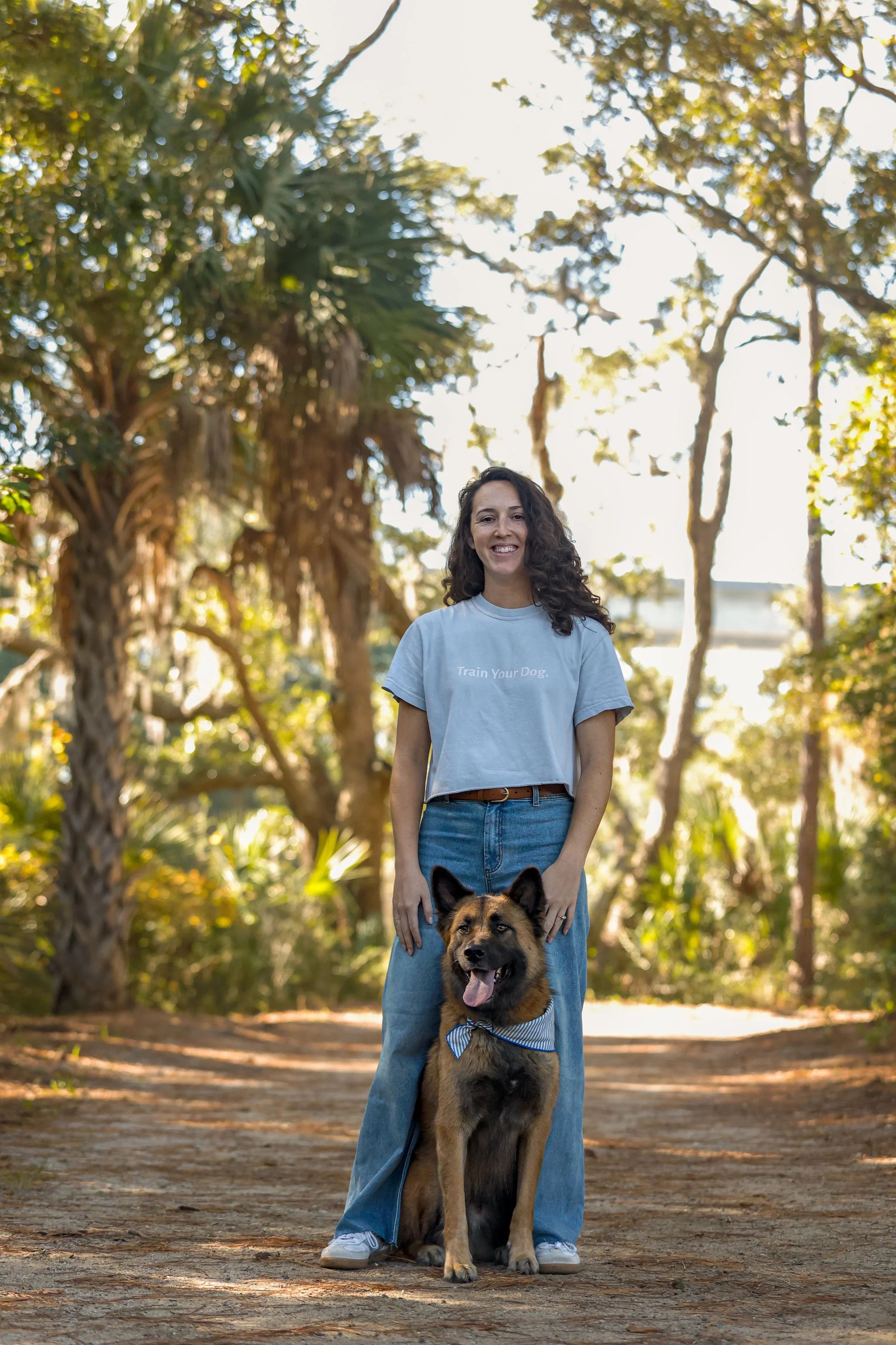 A woman standing outdoors on a dirt path with her dog. The woman has curly brown hair and is wearing a light gray T-shirt with the words "Train Your Dog" printed on it, along with blue jeans. The dog is a German Shepherd wearing a blue and white striped bandana, sitting in front of the woman. The background features tall trees and sunlight filtering through the leaves.