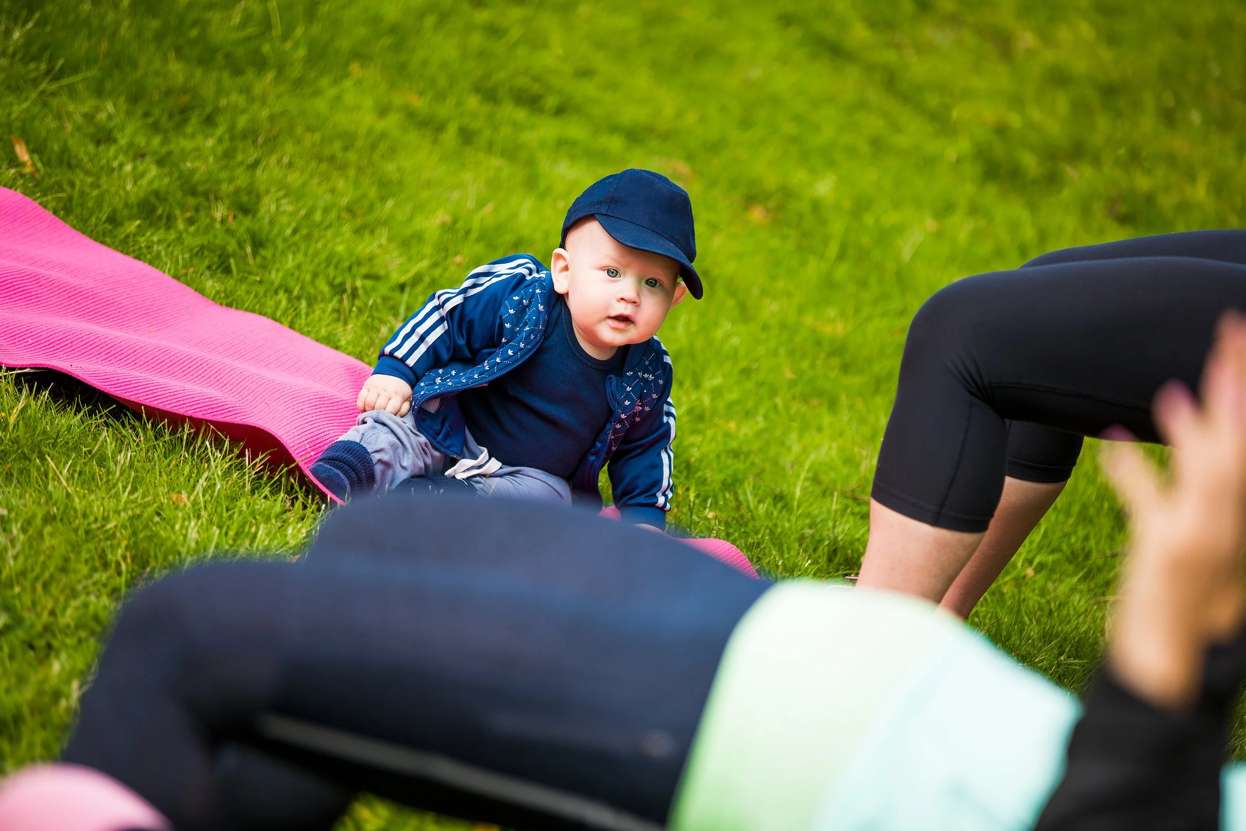 En bebis i blå kläder och mössa som ligger på magen på gräset under ett yoga- eller träningspass, omgiven av personer som deltar i liknande aktivitet.
