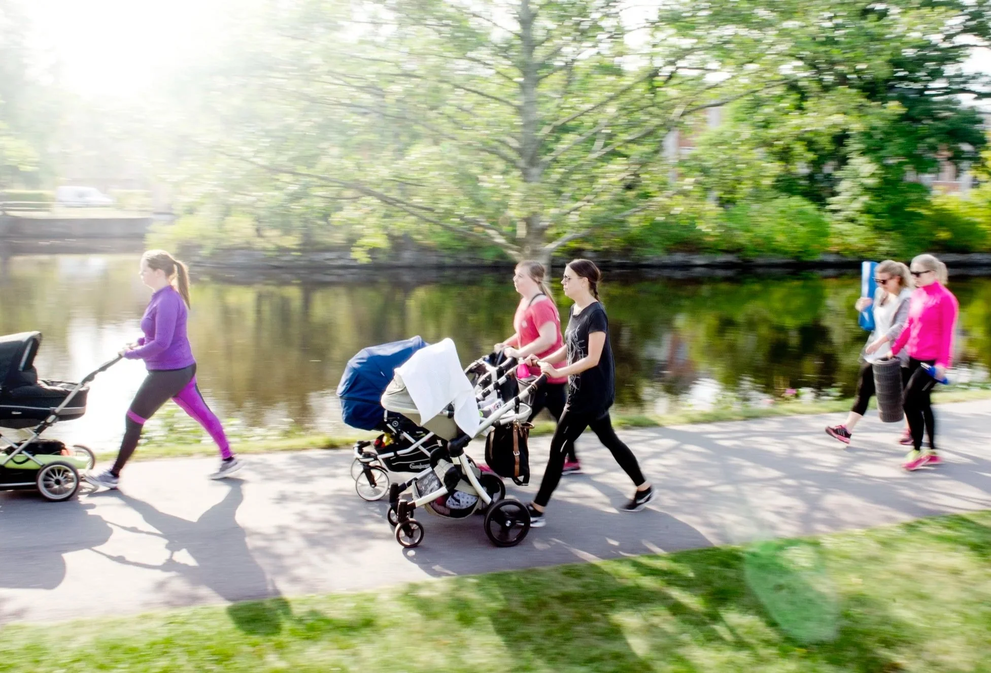 Flera personer, inklusive unga kvinnor, promenerar längs en sjöpromenad under en solig dag. De är på väg att gå förbi en trädinramad sjö.