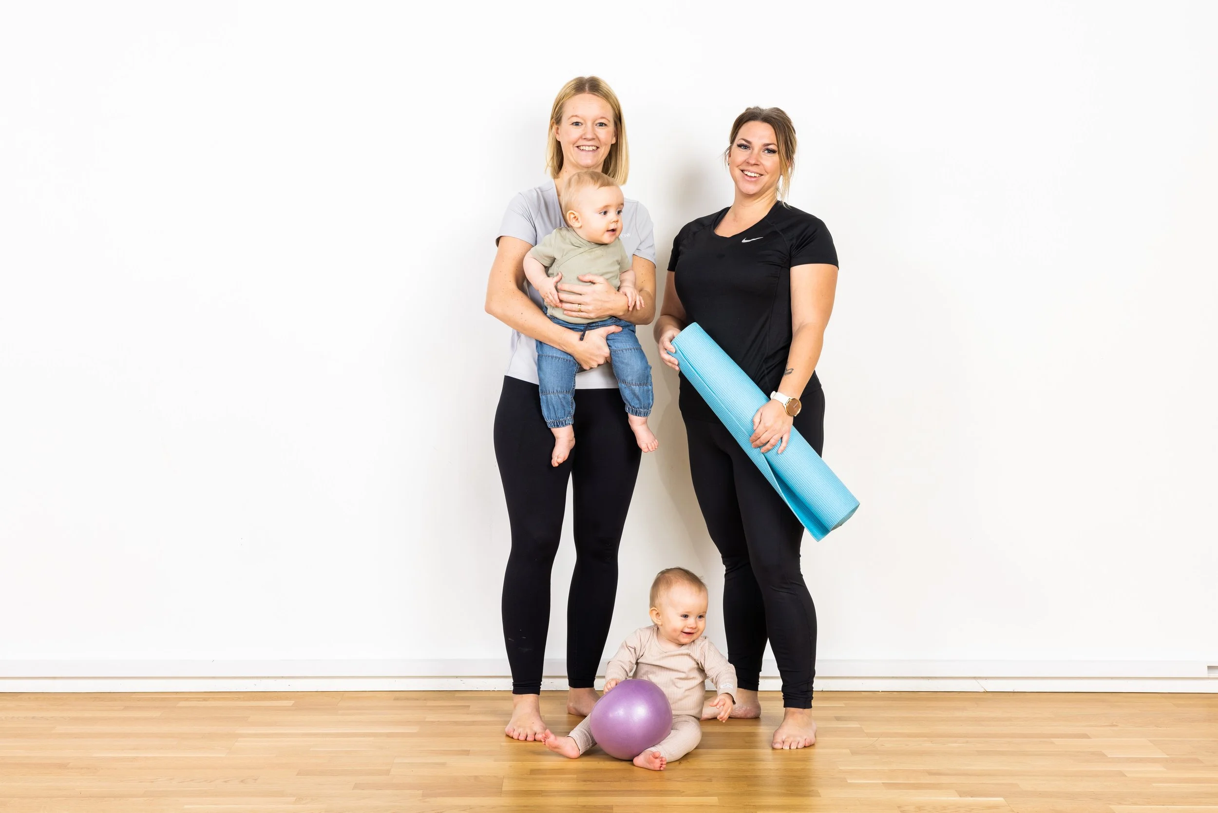 Två kvinnor och två barn, ett barn på golvet med en boll och ett annat som hålls av en kvinna, står framför en vit vägg, en av kvinnorna håller en blå yogamatta.