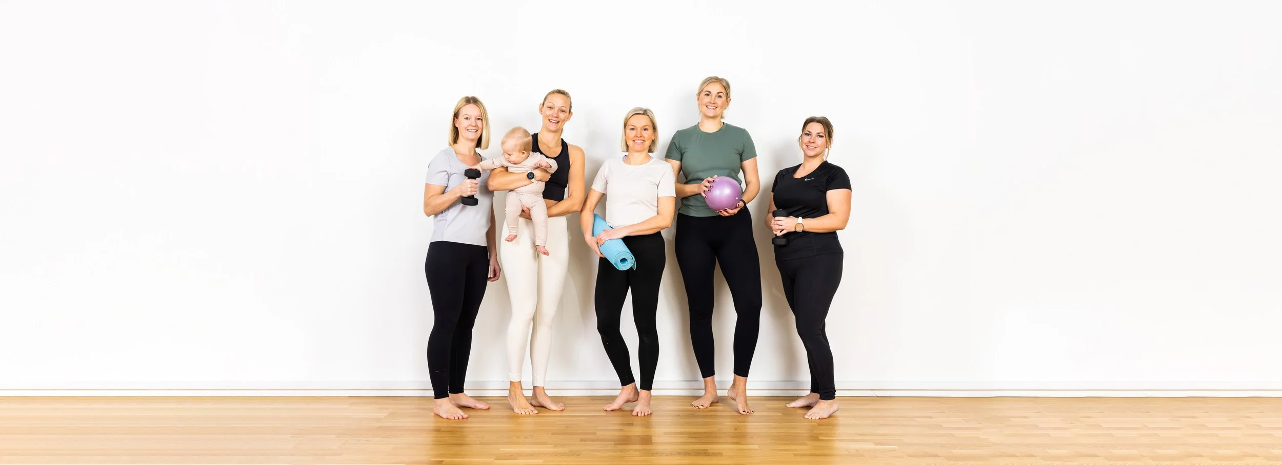 Grupp av kvinnor i träningskläder med ett barn, håller hantlar, pilatesboll och yogamatta i ett fitnesscenter.