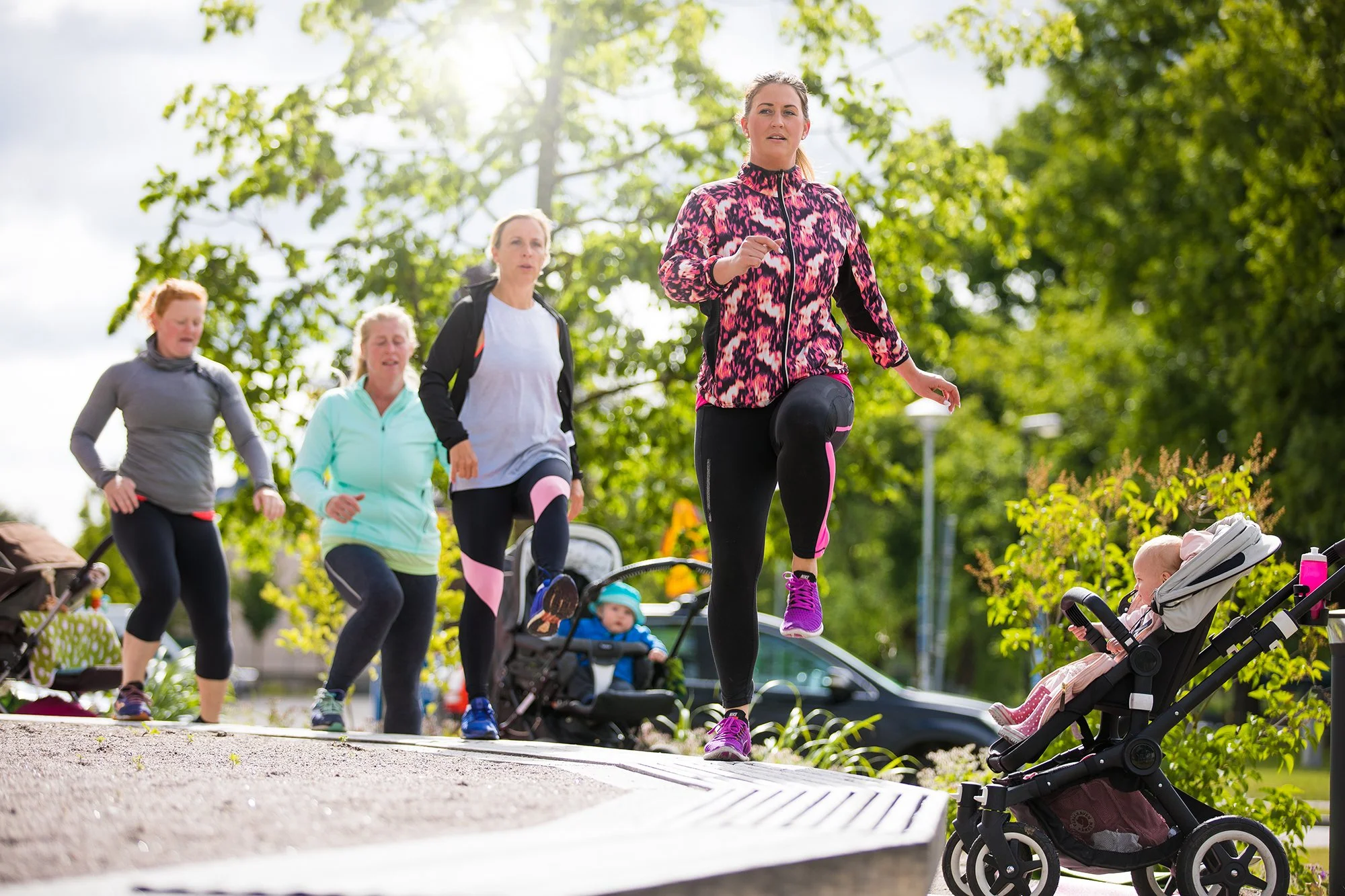Flera kvinnor och barn som deltar i en utomhus träningsklass i en park, med träd och gröna löv i bakgrunden, medan några barn sitter i barnvagnar.