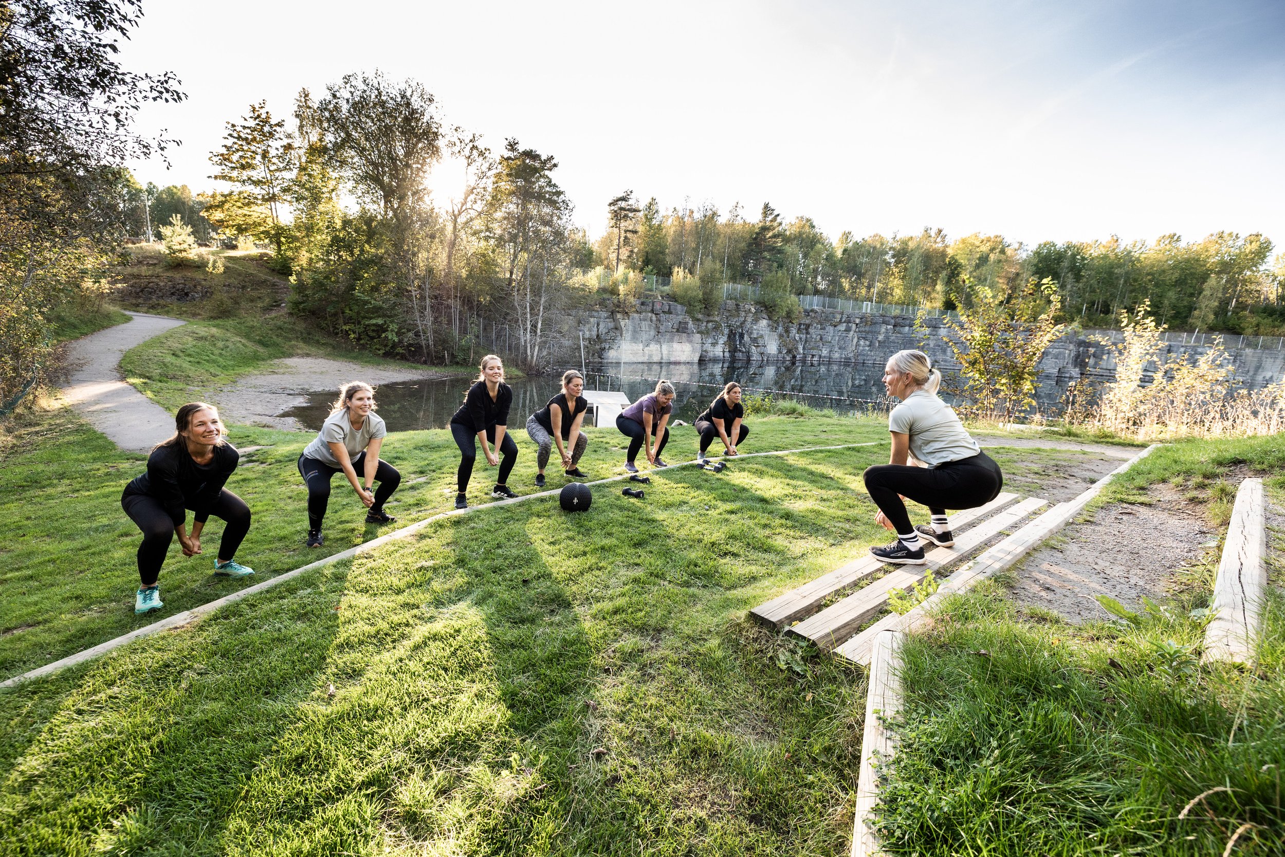 En grupp kvinnor utomhus tränar styrketräning och squats på gräsmattan vid en sjö i solen.