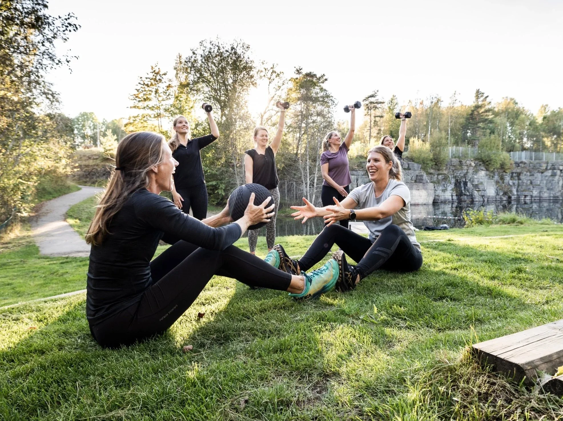 Grupp kvinnor som tränar utomhus, en kvinna hjälper en annan med en kettlebell medan andra gör armövningar.