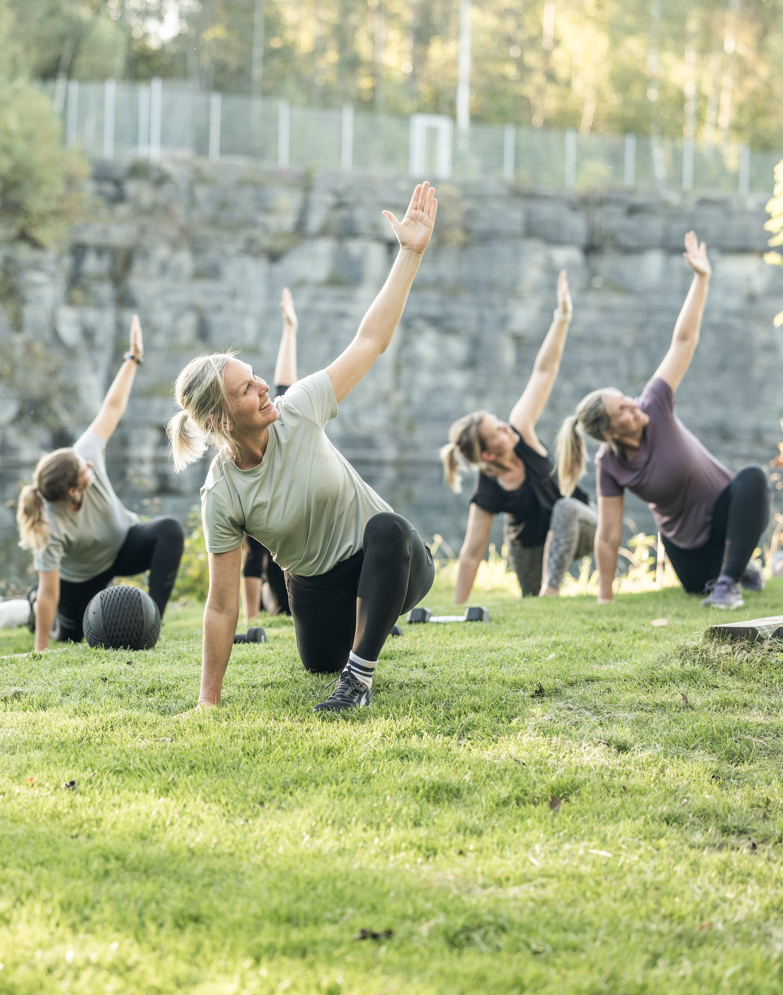 En grupp människor deltar i en utomhus yoga- eller stretchingsövning på gräset i en park, med träd och vatten i bakgrunden.