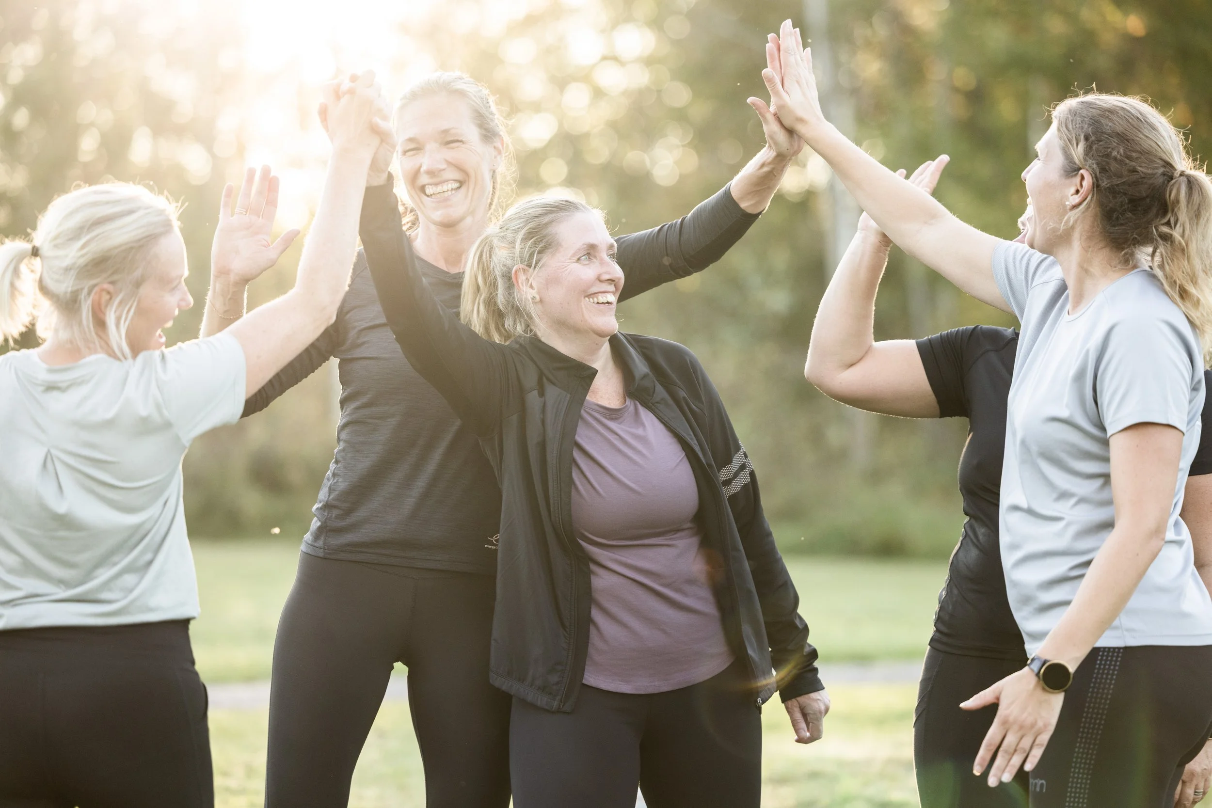 Fem kvinnor som är med i en grupp, high-five i en park under dagen.